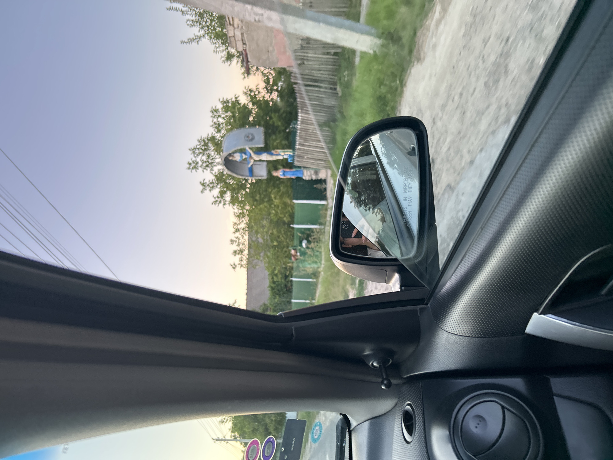 This is a first-person perspective photo taken from inside a vehicle, looking out the passenger side window. The image captures a rural scene, possibly in Abaclia, Moldova, during what appears to be either sunrise or sunset given the warm, soft lighting.
In the foreground, the interior of the car is visible, including the black textured door panel, a silver door handle, and a circular air vent. The side mirror reflects the interior of the car and the driver, who is wearing a white top and holding up a hand, possibly adjusting something or signaling. A warning sticker "OBJECTS IN MIRROR ARE CLOSER THAN THEY APPEAR" is present on the mirror's edge.
Outside the car, a dirt road or path is visible. On the right side of the frame, a weathered wooden fence runs along the edge of the road. Beyond the fence, there are green trees and some buildings, including a stone structure.
The focal point of the scene outside the car is a religious roadside shrine. It features a metal arch structure with a statue of Jesus Christ on the cross. To the left of the cross, a blue statue of a woman, likely the Virgin Mary, stands. The shrine is situated amongst trees and appears to be a common sight in rural areas of Eastern Europe. Power lines are visible stretching across the sky, suggesting the presence of infrastructure. The overall impression is one of a tranquil, everyday scene in a village or rural setting.