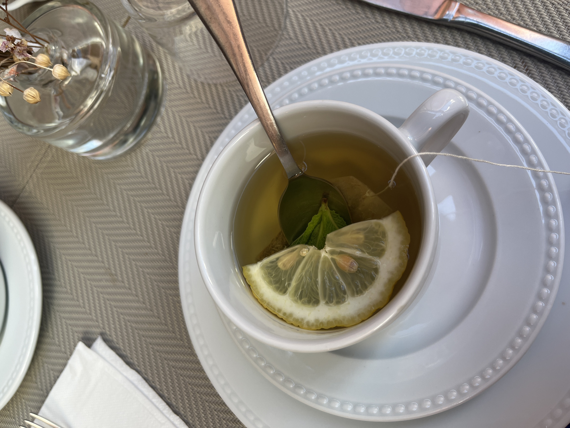 A close-up, high-angle shot shows a cup of tea with a lemon slice and mint leaves, set on a white saucer and plate with a beaded rim design, placed on a woven-textured, neutral-colored table covering. A teaspoon is present in the cup. A small glass vase with dried floral elements is visible in the background on the left side of the image. A white napkin, fork, and knife are positioned adjacent to the plate setting. The scene appears to be in Lisbon, Portugal.