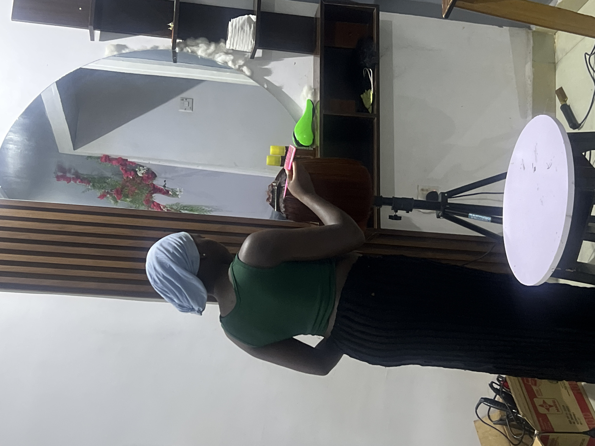 In Gura Top, Nigeria, a woman wearing a light blue headscarf and a green tank top stands near a mannequin head that is partially visible in the lower left of the frame. She is holding a pink comb and appears to be working on the mannequin's hair. The mannequin's hair is reddish-brown and styled in a bob. A tripod stands behind the mannequin. To the right of the mannequin, on a dark shelf, are three yellow bottles, a bright green hair dryer, and some white cotton-like material. Above the shelf, a large, oval mirror reflects a doorway with a floral arrangement of red and white flowers. The environment appears to be an indoor setting, possibly a salon or a room set up for hairstyling, with a white wall, wooden paneling, and a dark shelving unit. A white round stool is positioned in the foreground. A cardboard box filled with electrical cords and equipment is visible on the left side of the image.