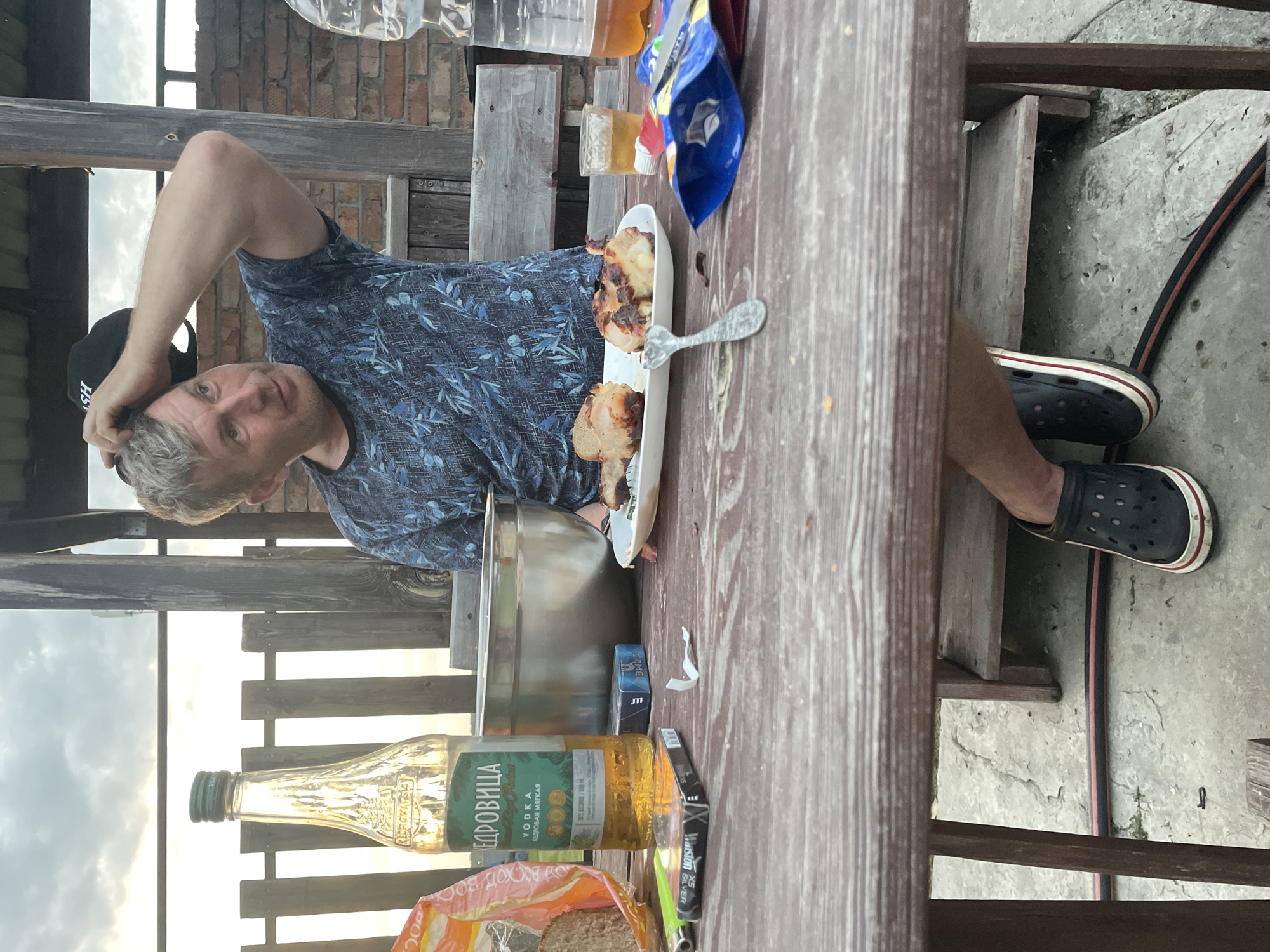 A middle-aged Caucasian man with graying hair and a blue patterned t-shirt is sitting at a wooden table outdoors, holding a black baseball cap on his head with his right hand. He is looking to his right. The table is covered with food and drinks, including a bottle of vodka labeled "КЕДРОВИЦА", a plate of grilled meat, a glass of amber liquid, a pack of cigarettes, and a bag of snacks. The background shows a wooden fence and a brick wall. The man is wearing black Crocs with a red and white trim. The scene appears to be a casual outdoor gathering, possibly in the city of Chulym, Russia, given the context.