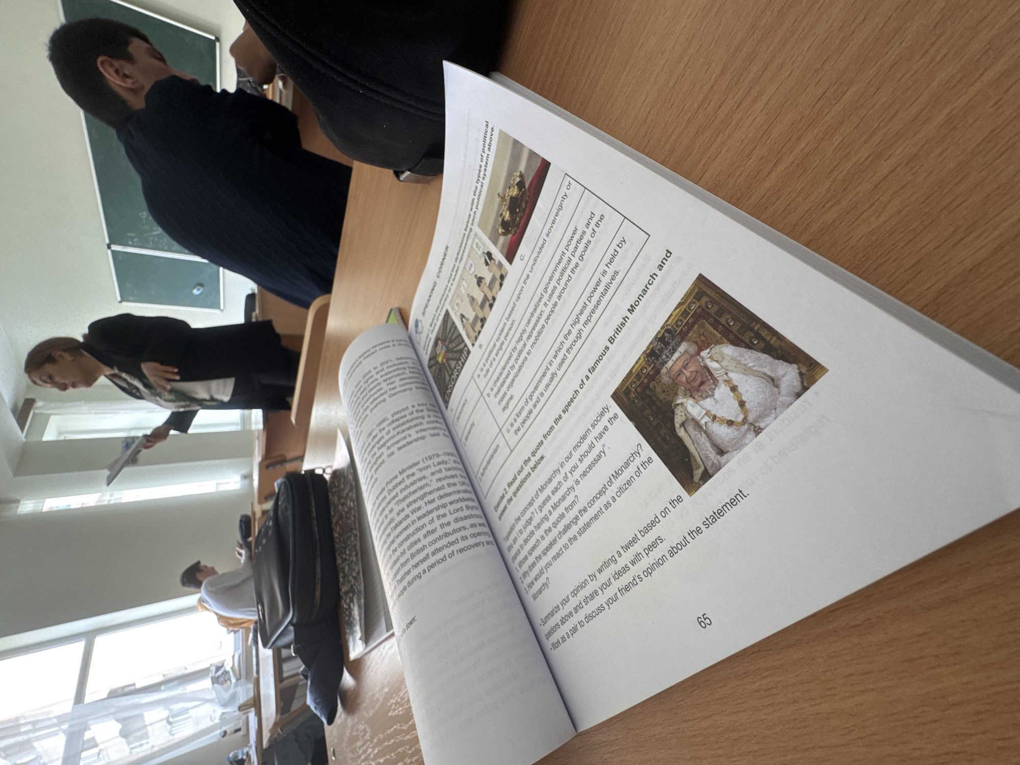 An indoor shot taken from a student's perspective, shows a classroom in Yerevan, Armenia during daytime, with natural light streaming through large windows on the left. In the immediate foreground, an open textbook rests on a light wooden desk. A black backpack is also visible on the desk to the left.
The textbook is open to page 65, displaying educational content. A prominent image shows Queen Elizabeth II in ceremonial attire. Adjacent text includes an exercise titled "Exercise 2. Read out the quote from the speech of a famous British Monarch and answer the questions below." The quote provided is: "1. I question the concept of Monarchy in our modern society. Who am I to judge? I guess each of you should have the chance to decide having a Monarchy is necessary'." Follow-up questions ask why the speaker challenges Monarchy and how a citizen would react. Further instructions prompt students to "Summarize your opinion by writing a tweet" and "Work as a pair to discuss your friend's opinion." Other visible text sections include "SPEAKING CORNER," "CENSORSHIP," "Authoritarianism," and definitions of different political systems, accompanied by small illustrative images like a crown. A section about Margaret Thatcher (Prime Minister 1979-1990) and the Falklands War is also partially visible on the left page.
Beyond the desk, a male student in a dark blue top is seated at another desk, seen from behind. Further back, a female teacher, dressed in a black jacket, stands near the windows, holding papers, seemingly addressing the class. Another student in a white top is visible further down the row of desks. A dark green blackboard is partially visible on the wall behind the first student. The overall scene depicts an active classroom environment.
