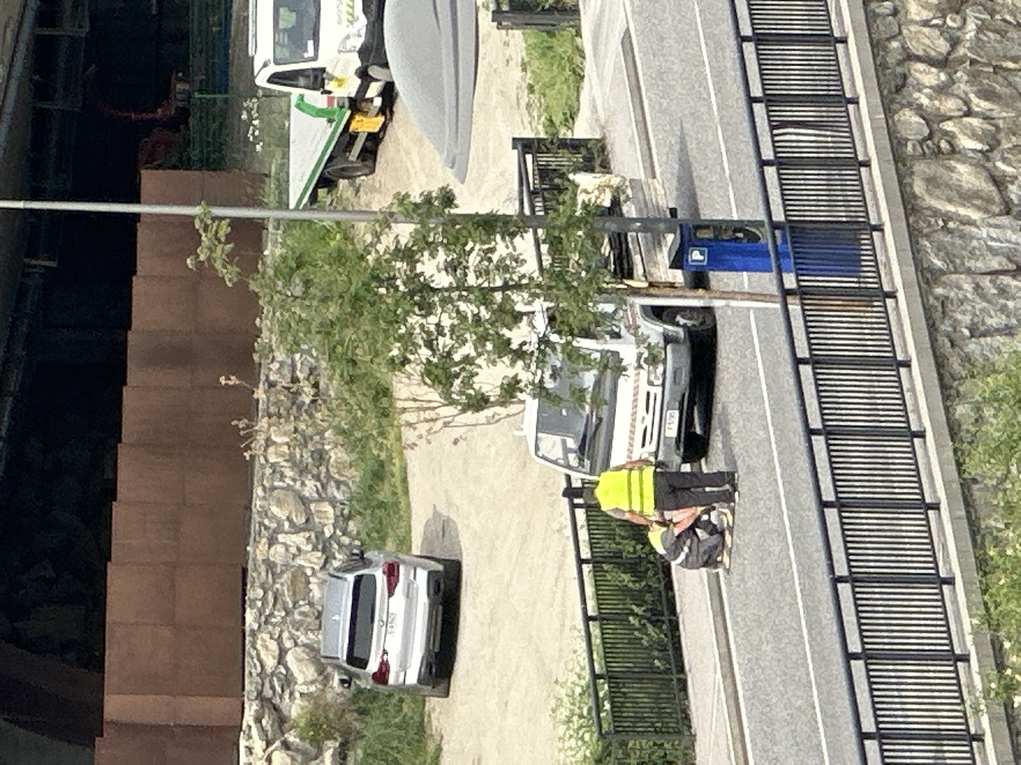 The image, rotated 90 degrees clockwise for proper viewing, captures an outdoor scene featuring a paved road, vehicles, and individuals, set against a backdrop of natural and man-made elements. The scene appears to be in a semi-urban or rural area, consistent with a mountain valley or town.
A two-lane paved road with white dividing lines runs horizontally across the mid-ground. To the left of the road, there is an unpaved dirt or gravel area, followed by a multi-story building constructed from dark brown, rectangular blocks. To the right of the road, a sturdy black metal fence or barrier, characterized by numerous horizontal slats, borders a rocky, vegetated embankment which includes a distinct dry stone wall.
Several vehicles are present. On the unpaved area to the left, a white pickup truck or utility vehicle, featuring green graphics and text like "K-500" on its side, is partially visible. On the paved road, further to the right, a white van is parked. Beside this van, a person dressed in a bright yellow high-visibility vest and dark trousers is seen bending over, seemingly engaged in work or inspection. Another individual or object is partially obscured next to them. Closer to the building on the paved road, a silver car, possibly a sedan or small SUV, is parked. Additionally, a blue utility box or small vehicle component with a white square bearing a 'P' symbol is visible near the black fence. The license plate "15765" is visible on the rear of the white van.
The scene is illuminated by bright daylight, suggesting clear weather. Green foliage from trees and bushes is scattered throughout the image, especially along the embankment. The overall impression is a candid shot of everyday activities, possibly maintenance or delivery work, unfolding in a location that blends natural landscape with infrastructural elements.
