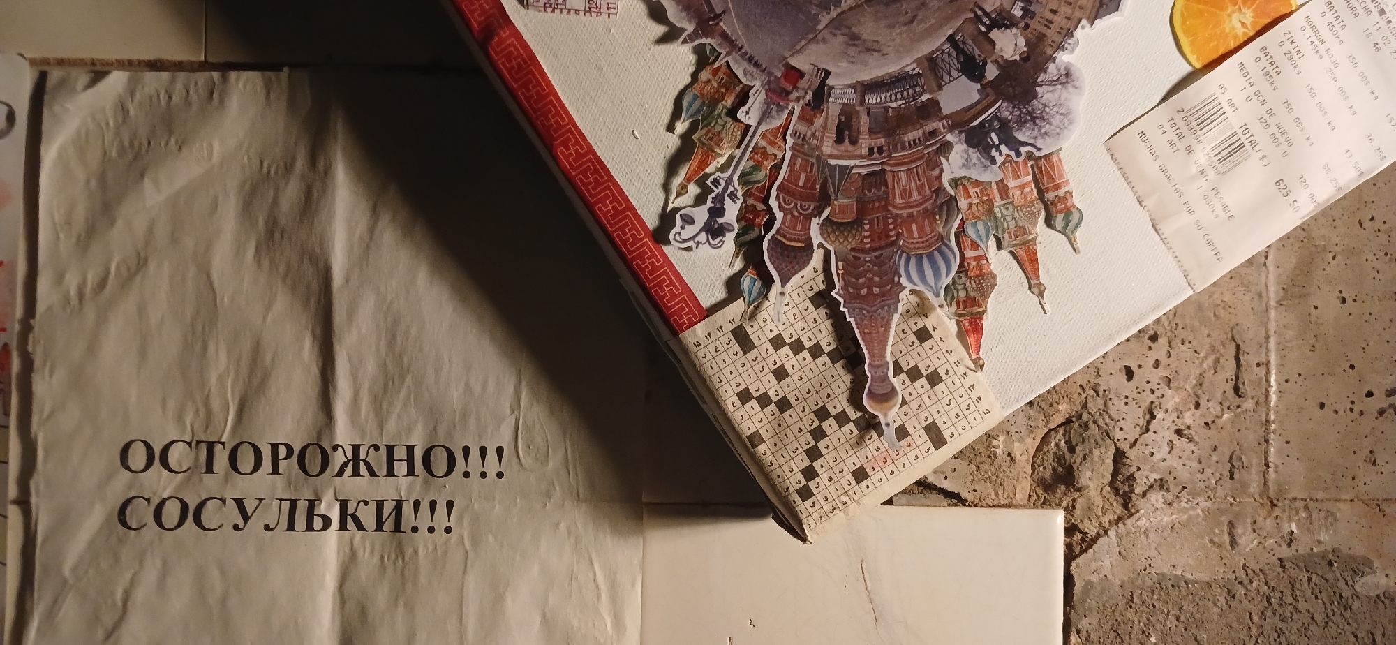 The media file displays a still-life composition featuring an eclectic mix of objects, hinting at cultural and personal themes.
The central element is a white board, possibly a canvas or piece of thick paper, adorned with a detailed paper collage. The collage consists of numerous cut-outs of architectural elements, primarily onion domes and elaborate facades, strongly resembling Moscow's iconic St. Basil's Cathedral. These elements are arranged in a spherical or "world" pattern. Below the collage, a section of a grid, possibly a crossword puzzle or game, with Arabic script is partially visible.
To the upper right, a crumpled supermarket receipt, likely from Argentina given the currency and Spanish phrases like "MUCHAS GRACIAS POR SU COMPRA" (Thank you for your purchase), lists items such as "BATATA," "MORRON ROJO," "ZIKINI," and "MEDIA DCN DE HUEVO" (half dozen eggs), totaling 625.50.
On the lower left, a crumpled beige paper displays a bold warning in Russian Cyrillic: "ОСТОРОЖНО!!!" (CAUTION!!!) and "СОСУЛЬКИ!!!" (ICICLES!!!). This is a common public safety announcement in Russia during winter.
The objects are arranged on a rough, textured surface, likely concrete or stone, with a clean white tile or object visible in the bottom right corner. The lighting is somewhat dim, creating soft shadows. The juxtaposition of Russian cultural imagery and warnings with an Argentinian receipt suggests themes of travel, memory, or multicultural connections.