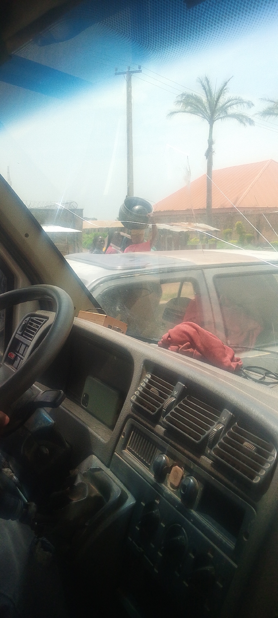 The image is an interior view of a vehicle, looking through the windshield. The driver's side steering wheel and dashboard are in the foreground. The dashboard has an air vent, a slot with a green smartphone, and other controls. Outside the vehicle, through the windshield, we see a street scene. A utility pole with wires is visible, along with a palm tree and a building with an orange roof. Several people are seen standing on the roof of another vehicle in the distance. One person is carrying a large black pot on their head. The sky is bright, indicating daytime. Based on context, the image is located in Kwang, Nigeria.