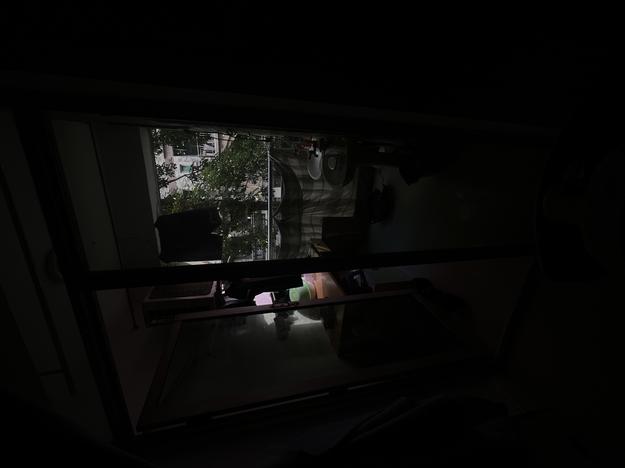 The image depicts a view through a glass door into a cluttered interior and a balcony area. In the dimly lit interior, an air conditioning unit is visible mounted on a wall. A table with various objects, including what appears to be a green hat, is also present. On the balcony, two sinks with faucets are installed. A drying rack with clothes hangs behind the sinks, and a patterned cloth is draped over a railing. Green trees and a building with windows are visible beyond the balcony. The overall scene suggests a domestic environment, possibly in an urban setting like Yuzhong District, China. The lighting suggests it is either early morning or late evening.