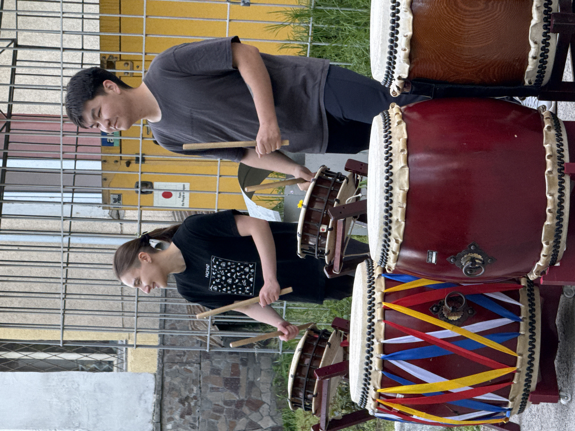Two individuals, one man and one woman, are playing taiko drums. The man is wearing a grey t-shirt and dark pants, and the woman is wearing a black t-shirt with a design on it and dark pants. They are both holding drumsticks. There are three taiko drums visible, stacked on top of each other. The bottom drum is decorated with red, white, blue, and yellow ribbons. The middle drum is red, and the top drum is brown. The scene appears to be taking place outdoors, possibly in the city of Bishkek City, Kyrgyzstan. A yellow building and a metal fence are visible in the background.