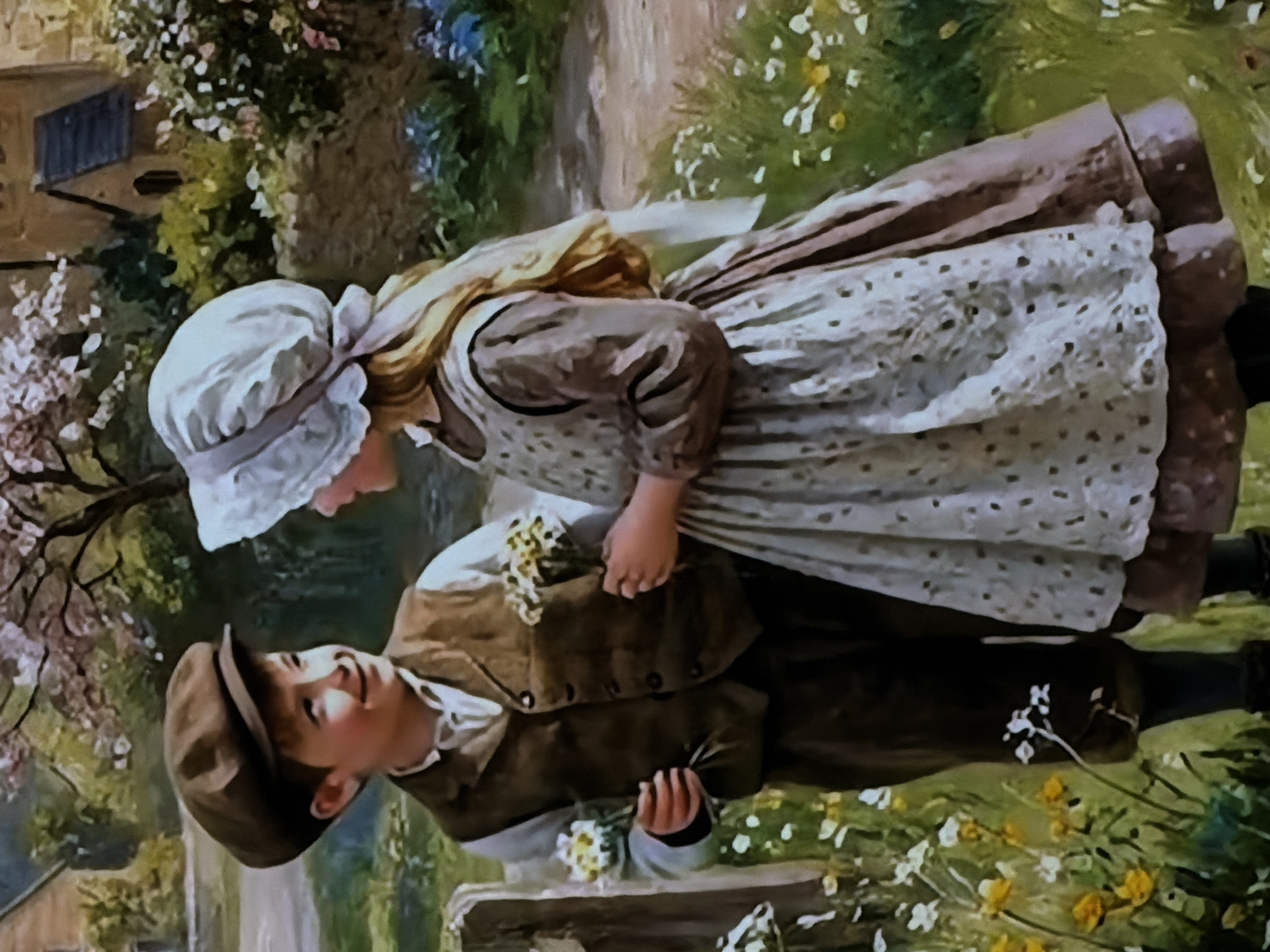 This image is a painting depicting two children in a lush, outdoor garden or rural setting. The painting is presented with a vertical orientation, as if rotated 90 degrees clockwise from its original landscape or portrait presentation.
The main subjects are a young girl and a younger boy, positioned closely to each other. The girl stands on the left, looking down at the boy with a gentle expression. She has light-colored hair, worn long, and is dressed in a white bonnet tied with a ribbon, a long-sleeved dark dress, and a light-colored, patterned or polka-dotted apron over it. She holds a small cluster of white flowers in her left hand, which is close to the boy's right arm.
The boy, positioned on the right and slightly lower, looks up at the girl with a clear smile on his face. He wears a brown flat cap, a white shirt, and a brown waistcoat or jacket with visible buttons, along with dark trousers. In his left hand, he holds a small bouquet of white and yellow flowers. He appears to be leaning against or sitting on a low stone wall or bench, which is partially visible at the bottom of the frame.
The background is rich with greenery, including trees, flowering bushes with white and pink blossoms, and patches of yellow and white wildflowers on the ground. A dirt path is visible behind the children, winding through the foliage. In the upper left background, part of a rustic building with a dark window or door can be seen.
The scene conveys a sense of innocence, tenderness, and youthful charm, set on a bright, sunny day, likely in spring or summer. The overall style is reminiscent of romantic or pastoral art, with a focus on natural light and delicate details. There is no visible text in the painting.
The location context for this media file is Sofiivska Borshchahivka, Ukraine.