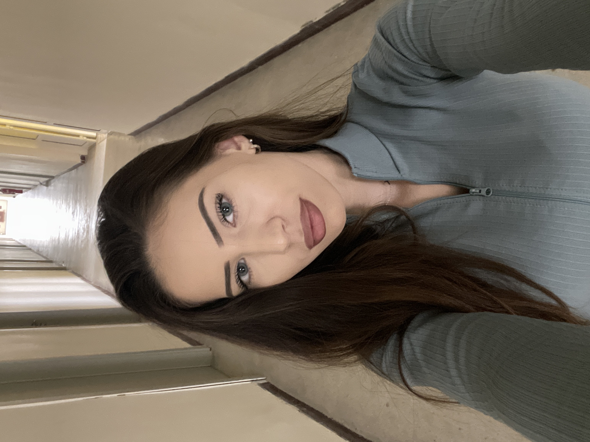 A selfie photograph depicts a young woman with long brown hair, light blue eyes, and fair skin, wearing a light blue zipper-front top. She is positioned in a hallway setting, likely indoors based on the artificial lighting and enclosed space. The hallway has light-colored walls and a tiled floor. The background suggests a building interior, possibly an institutional or public space, with architectural details suggestive of an older structure. There are no definitive elements to pinpoint the location to Veliko Tarnovo, Bulgaria.