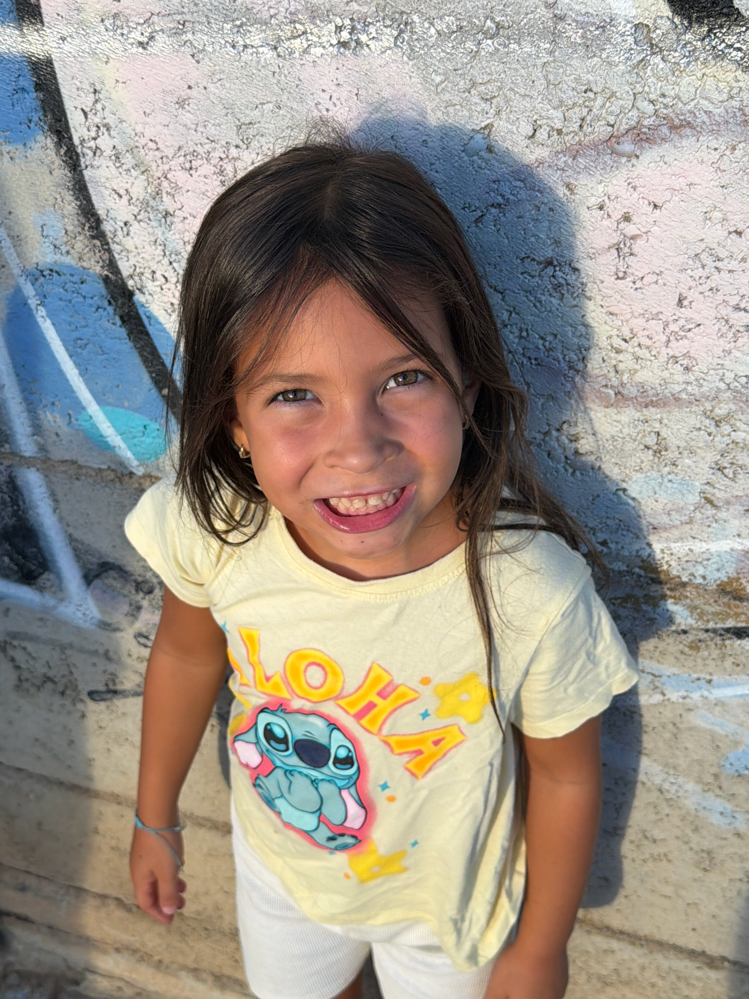 A young girl with long dark hair and brown eyes smiles directly at the camera. She is wearing a pale yellow t-shirt with a graphic of the character Stitch and the word "ALOHA" printed on it, and white shorts. A thin blue bracelet is visible on her left wrist. She is standing against a textured, light-colored wall with graffiti on it. The lighting suggests it is daytime, with a shadow cast on the wall from the left. The scene appears to be outdoors.