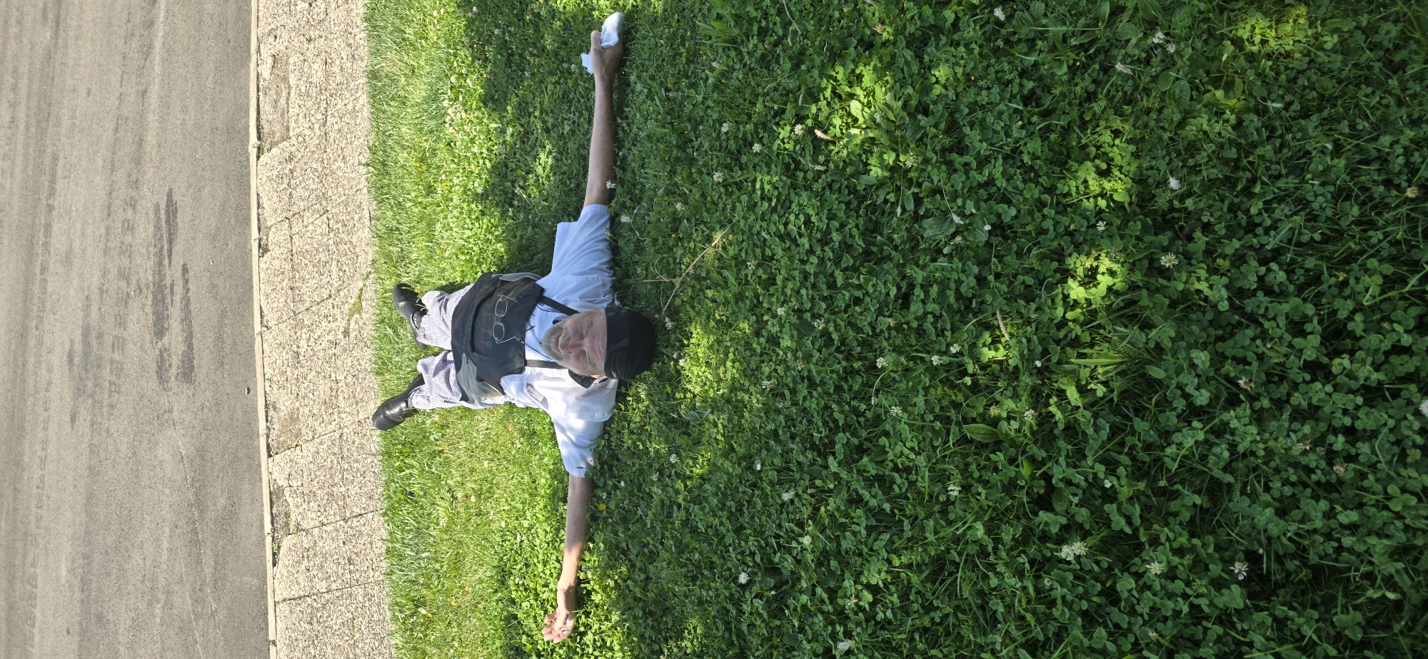 An individual is lying on their back in a grassy area next to a paved sidewalk and a road in Sallent de Gállego, Spain. The person is wearing a white collared shirt, striped pants, black shoes, and a dark vest with suspenders. They have a black head covering and eyeglasses perched on their chest. Their arms are extended outwards, and their right hand holds a white object, possibly a tissue or cloth. The grass is green and appears to be a mixture of grass and clover with some small white flowers. The sun is shining, casting shadows on the grass. The pavement in the background shows signs of wear.
