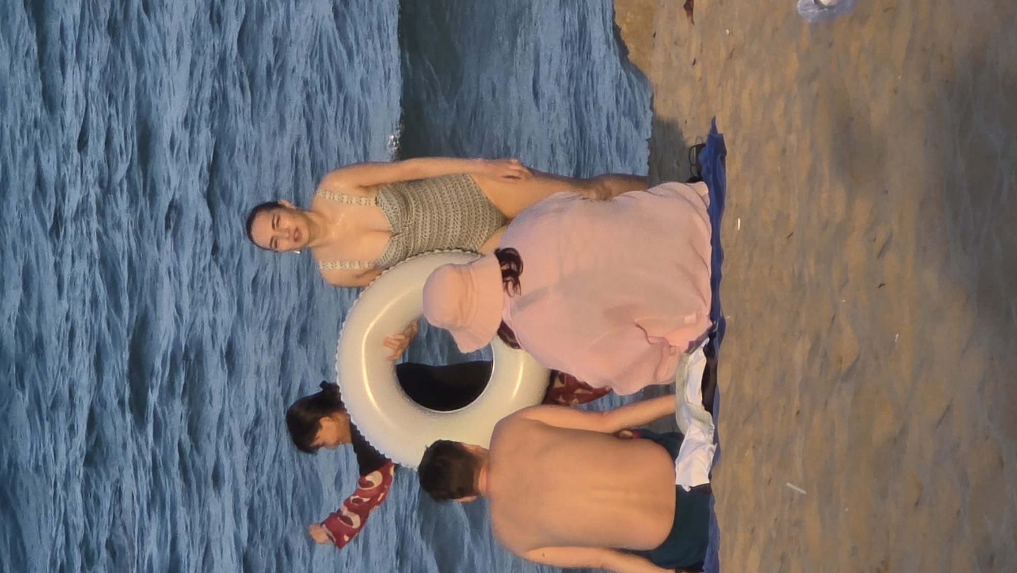 The image shows four individuals at a beach in Nha Trang, Vietnam. One woman is in the water wearing a knitted bathing suit, holding onto a white inflatable ring. Another individual, wearing a pink outfit with a hat, is sitting on the sand. A shirtless man is also on the beach with what appears to be dark swim trunks. Part of a dark-haired person wearing a red patterned shirt and glasses is visible. The beach is sandy and next to the ocean. A towel lies on the sand next to them.