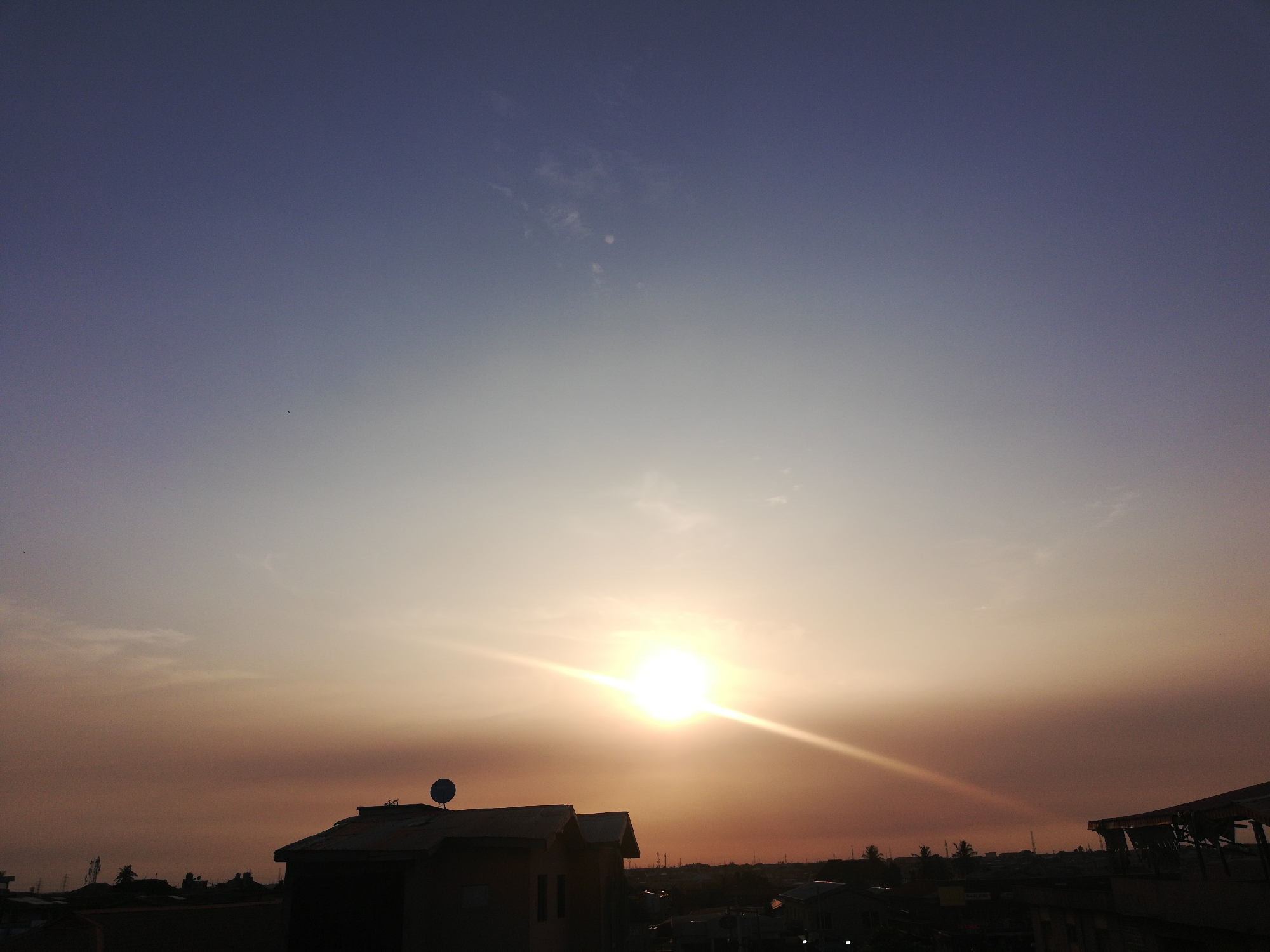 This image captures a vibrant sunset or sunrise over a semi-urban landscape in Lagos, Nigeria. The sky dominates the frame, showcasing a striking gradient of colors. The upper portion is a deep, muted blue and purple, transitioning downwards to a pale, ethereal blue-grey, and finally into brilliant oranges and yellows around the horizon. The sun, a radiant, almost white orb, is positioned slightly to the right of the center, emitting intense light and strong lens flares that stretch horizontally across the sky.
Below the horizon, the silhouettes of numerous buildings, rooftops, and some indistinct vegetation are visible. These structures are heavily shadowed, appearing as dark masses against the brighter sky. On the roof of a prominent building on the left side of the frame, a distinct satellite dish is silhouetted. The overall scene suggests a peaceful moment at either dawn or dusk, characterized by warm, soft light and atmospheric haze. There are faint, wispy clouds scattered across the sky. No people or visible text are present in the image.
