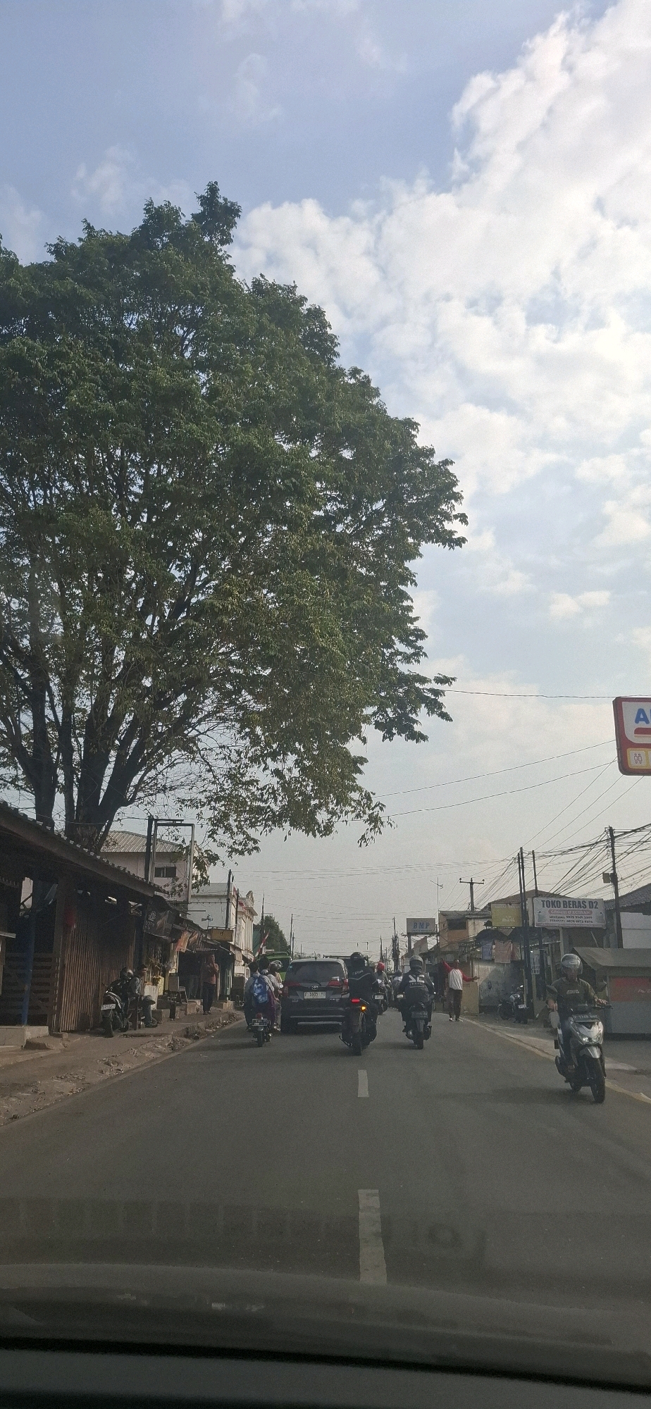 The image depicts a street scene in Sirnagalih, Indonesia, under a partly cloudy sky. A large, leafy tree dominates the left side of the frame, casting a shadow over a row of low-rise buildings that appear to be shops or stalls. The buildings have corrugated metal roofs and wooden fronts, some with open doorways.
The street is paved and shows signs of traffic. Several motorcycles are visible, some with riders, traveling in the same direction as the camera. A dark SUV is also present in the traffic. One rider is wearing a black jacket with white lettering. On the sidewalk to the left, several people are standing or sitting near motorcycles. One person is wearing a white shirt and red pants, gesturing with their arms.
To the right of the street, there are more buildings, including one with a sign that reads "TOKO BERAS DJ." Power lines crisscross the sky. The overall impression is of a busy, functional street in a tropical urban or semi-urban environment.