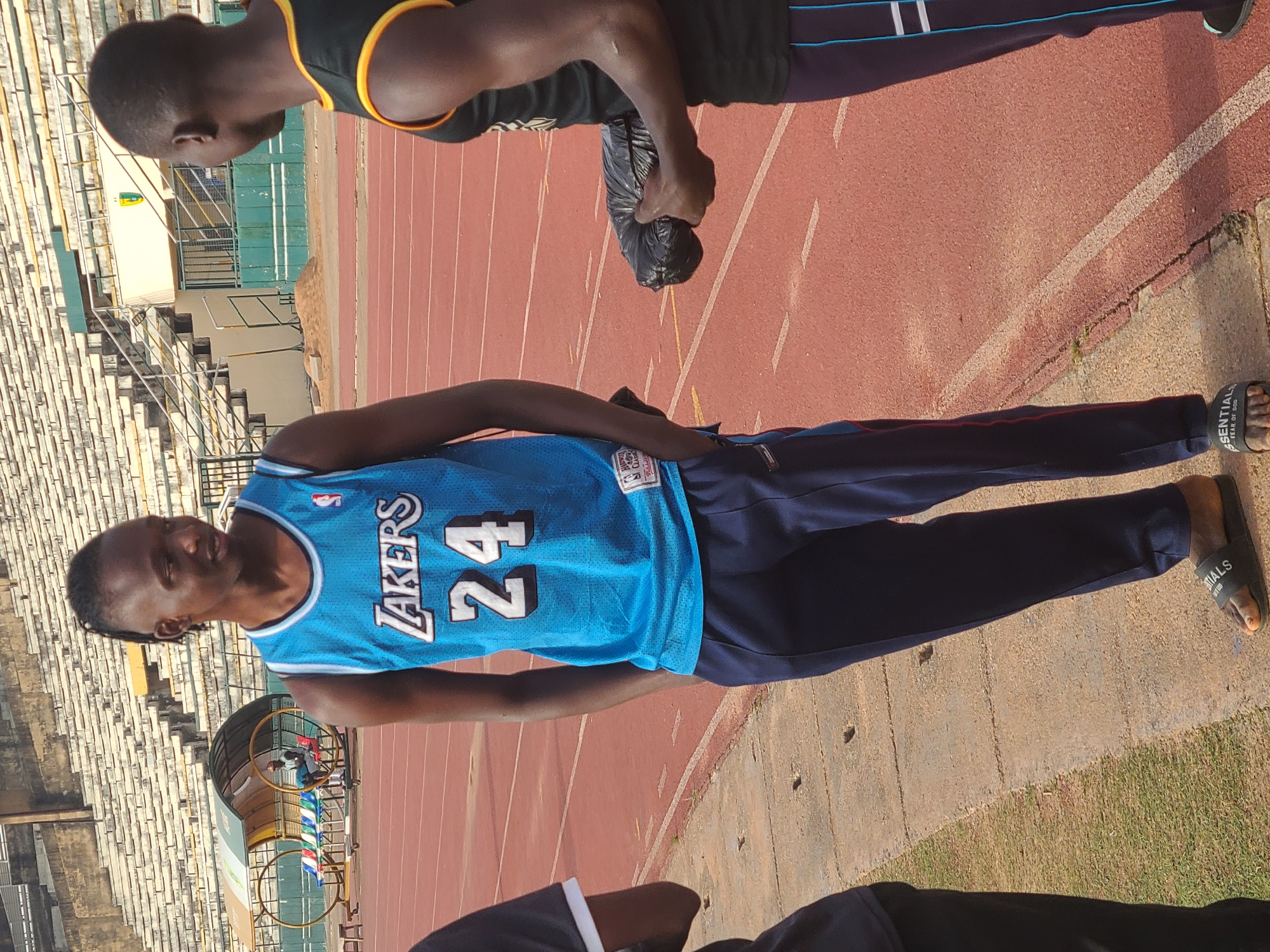 The image shows a man standing on a track at what appears to be a stadium. He is wearing a blue Lakers jersey with the number 24. He is also wearing dark-colored pants and slides. Another person is partially visible in the top of the frame, wearing a dark tank top with yellow trim. The track is red, and there are stadium seats in the background. Based on the information provided, the location is New Stadium, Nigeria.