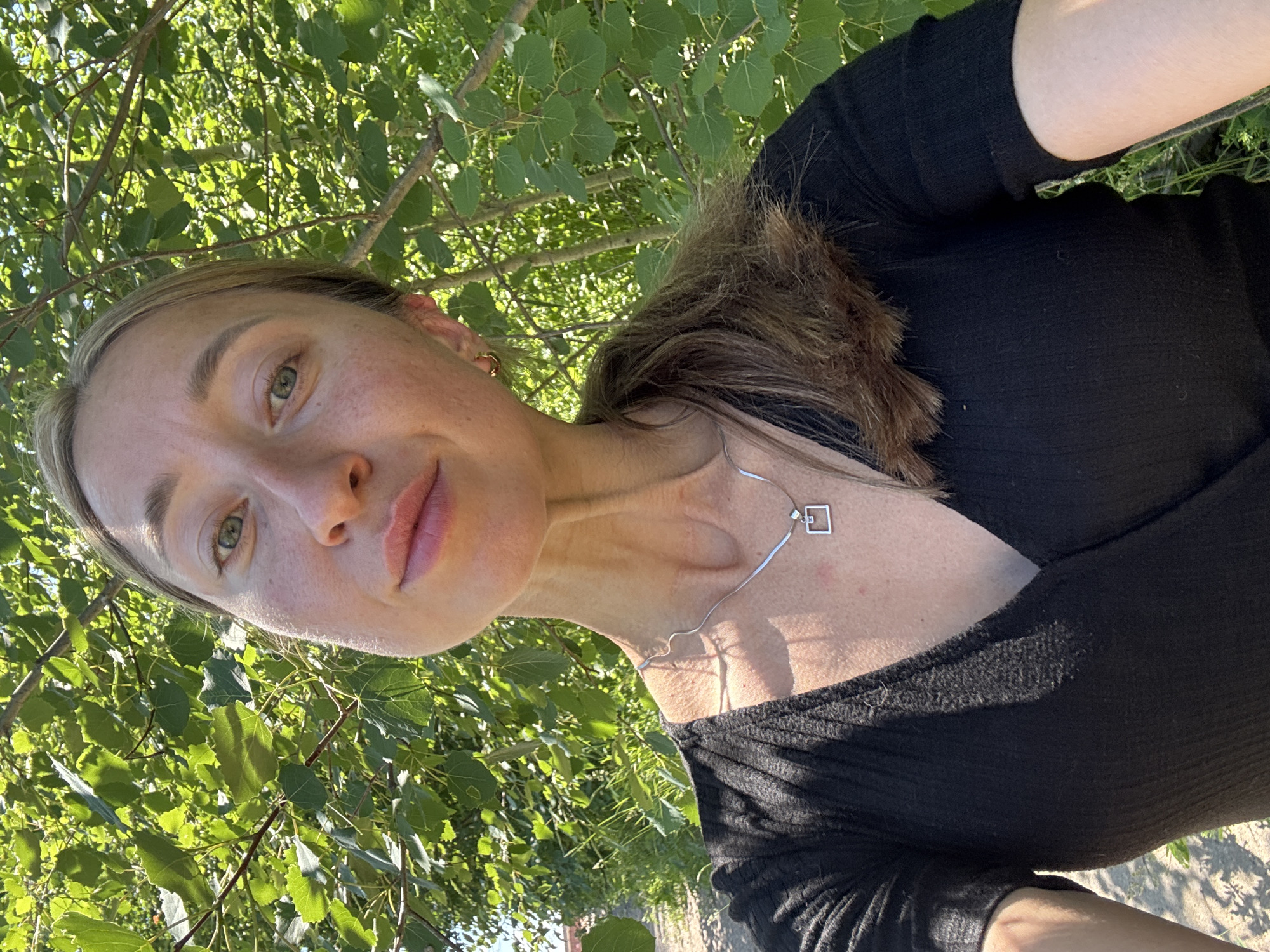 A woman with light brown hair and fair skin is taking a selfie outdoors, surrounded by lush green foliage. She is wearing a black, v-neck top and a silver necklace with a small square pendant. Sunlight filters through the leaves, creating dappled light on her face and clothing. The background consists of trees with vibrant green leaves and visible branches. The overall setting appears to be a natural, possibly park-like, environment within the city of Chaikovsky, Russia.