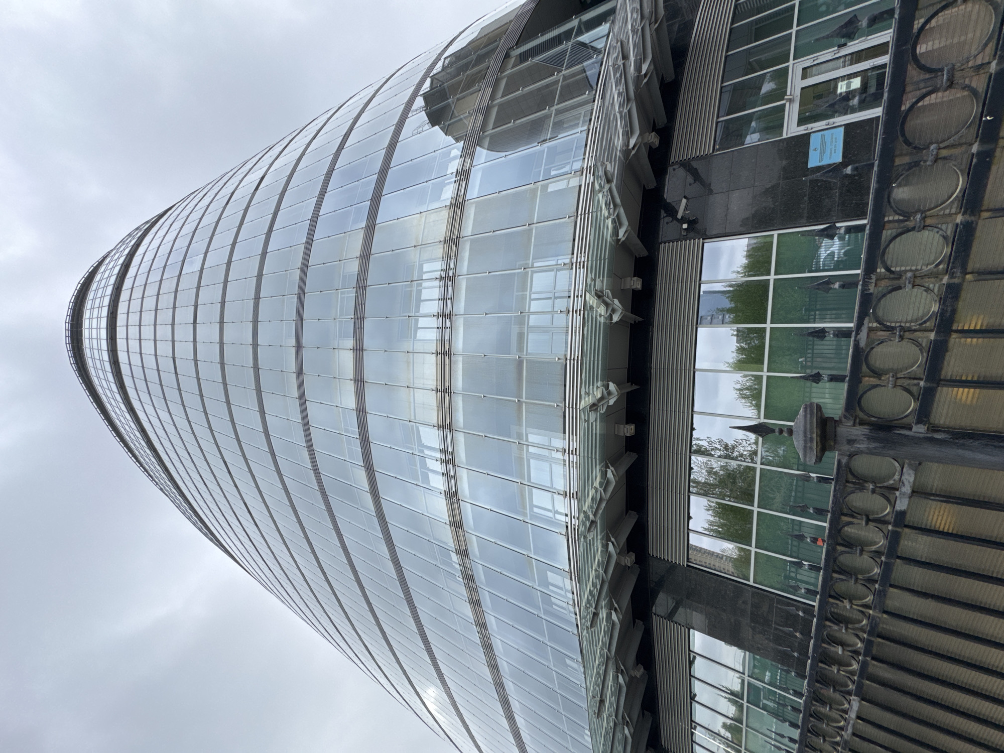 The image presents a low-angle view of a cylindrical glass-fronted building in Saint Petersburg, Russia, with a distinctive, curved architectural design. The building's facade comprises multiple glass panels, giving it a sleek, modern appearance. The cloudy sky reflects off the glass, creating an interplay of light and shadow across the surface. Adjacent to the main structure is a section with a dark, possibly granite or marble, finish that features barred windows and what seems to be an ornately decorated metal support structure. The overall environment is urban and likely located in a business or commercial district within Saint Petersburg.