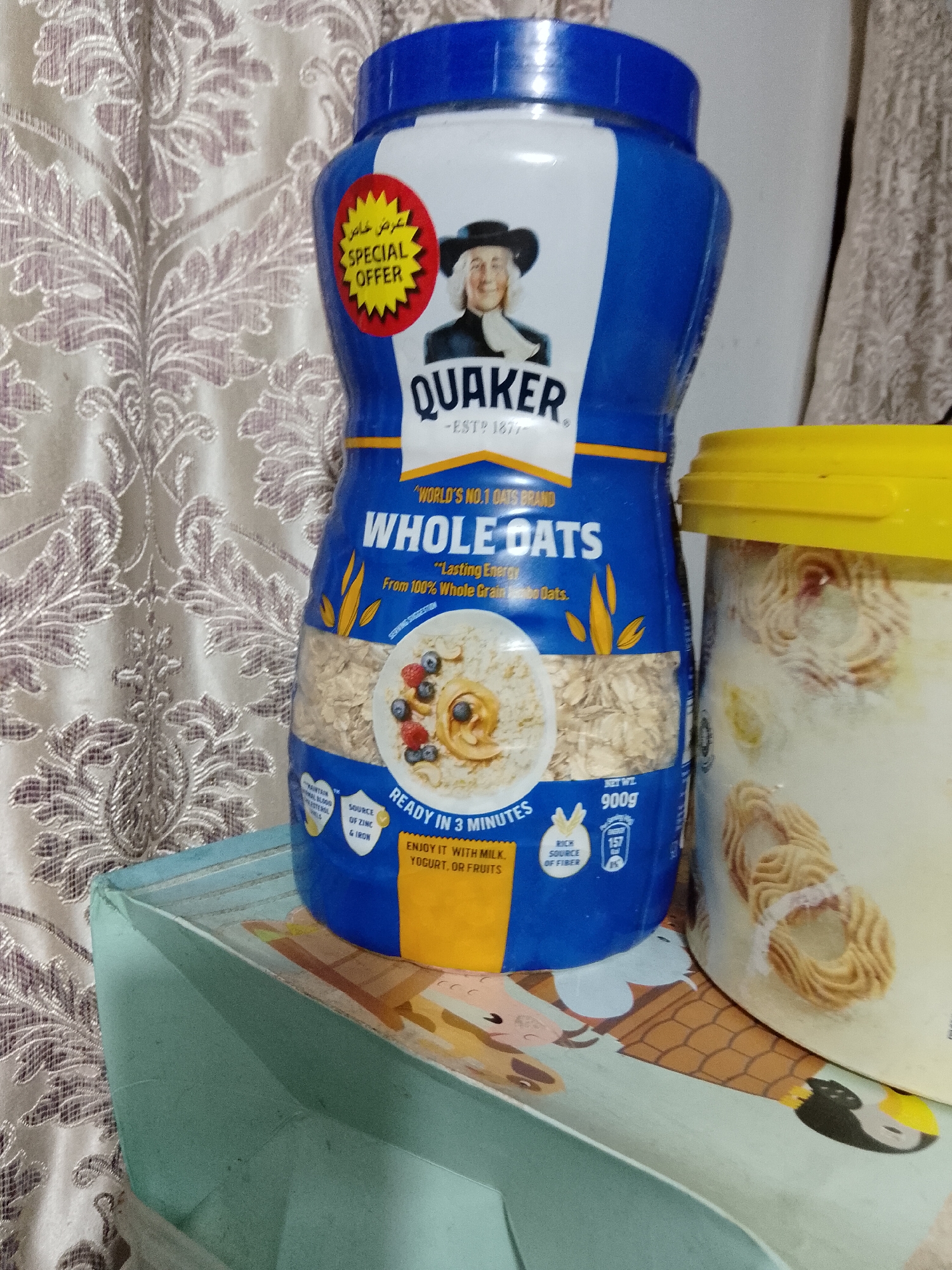 The image displays a still life composition featuring two food containers on a light blue surface, set against a patterned curtain.
The primary subject is a blue and white Quaker Oats container, prominently positioned on the left. It features the brand's iconic Quaker man logo. A red and yellow starburst sticker on the container reads "SPECIAL OFFER" in both English and Arabic ("عرض خاص"). Visible text on the label includes "QUAKER," "EST? 1877," "WORLD'S NO.1 OATS BRAND," and "WHOLE OATS." Further details state "Lasting Energy" "From 100% Whole Grain Oats," "READY IN 3 MINUTES," and suggests to "ENJOY IT WITH MILK, YOGURT, OR FRUITS." It also highlights being a "SOURCE OF ZINC & IRON" and "RICH SOURCE OF FIBER." The net weight is indicated as "NET WT. 900g." An illustration of a bowl of oats with berries and yogurt is also visible on the label.
To the right, partially visible, is a white plastic container with a bright yellow lid, which appears to hold numerous light-colored, ring-shaped cookies or biscuits.
The background is a patterned curtain featuring an ornate, repeating floral or paisley design in muted, possibly beige or light grey, tones. The light blue surface supporting the containers appears to be a tray or box.
The scene is a domestic setting, likely indoors in Davao City, Philippines, given the provided context. No specific activity or event is taking place, and the time of day or emotional context is not discernible.
