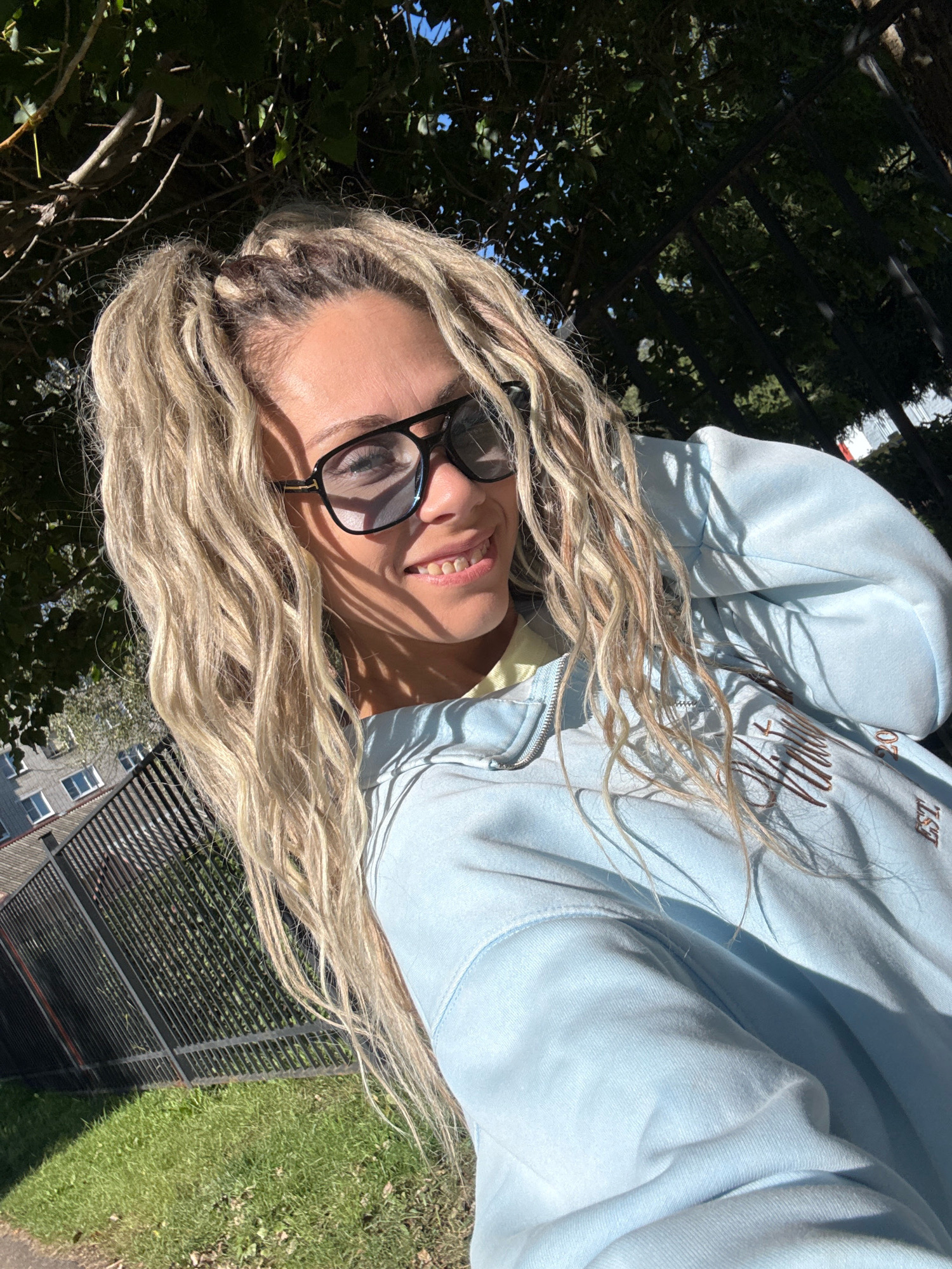 A woman with blonde dreadlocks is taking a selfie outdoors. She is wearing black-framed glasses and a light blue zip-up hoodie. The background includes trees, a black metal fence, a grassy area, and a multi-story building with windows. The lighting suggests it is daytime, with sunlight casting shadows. The scene appears to be in an urban or suburban environment, possibly in Zhukovo, Russia, given the architectural style of the building and the type of fence.