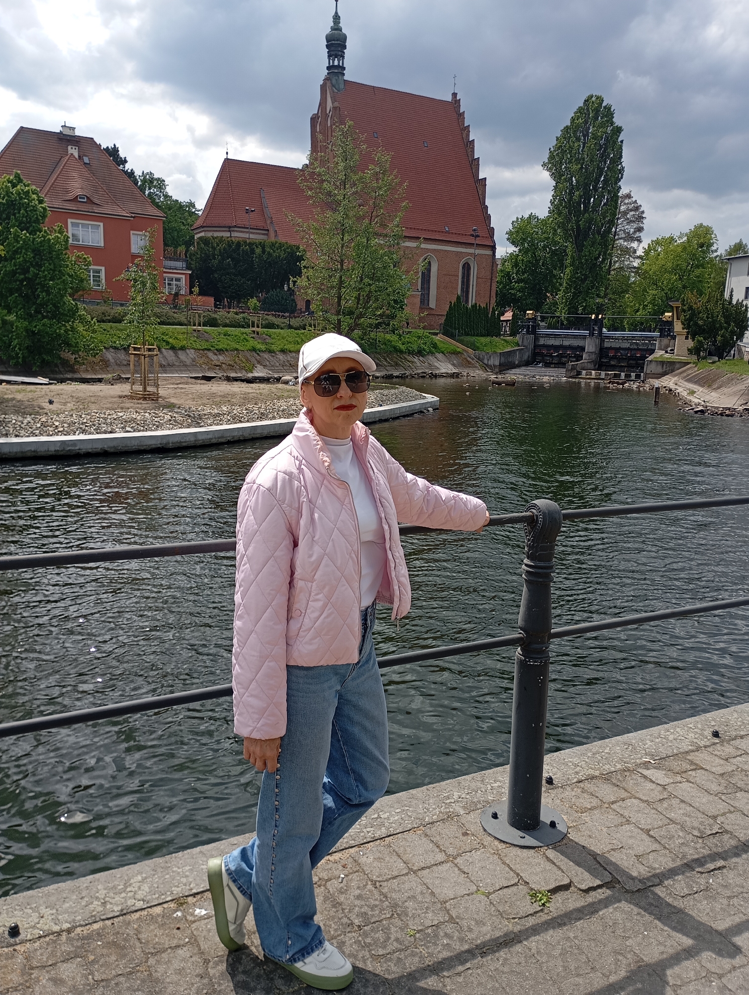 A full outdoor shot shows a woman standing next to the Brda River in Bydgoszcz, Poland. She is leaning on a black metal railing. She is wearing sunglasses, a white baseball cap, a pink quilted jacket, a white t-shirt, blue jeans with a row of silver-colored studs down the side seam, and white and light-green tennis shoes.
Behind her and across the river are a red-brick building with a tall steeple, a red building, and trees. To the right of the steeple is a dam. The weather is cloudy. The ground is paved.