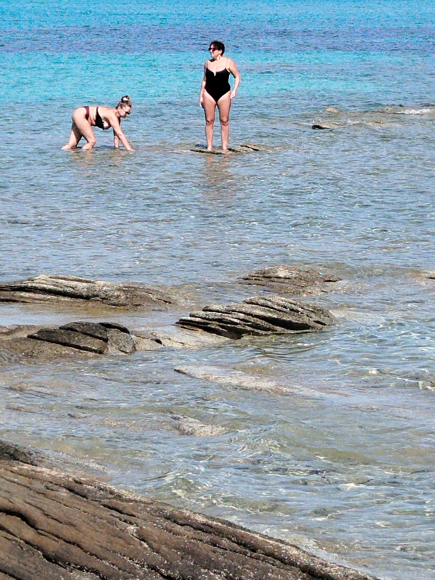 Two women are standing in shallow, clear turquoise water near a rocky shore. The woman on the left is wearing a black bikini with a maroon trim and is on her hands and knees. The woman on the right is wearing a black one-piece swimsuit and is standing upright. The water is calm with small ripples. The sun is shining, creating a bright and serene atmosphere. The location appears to be a beach in Sithonia Municipal Unit, Greece, known for its clear waters and scenic beauty.