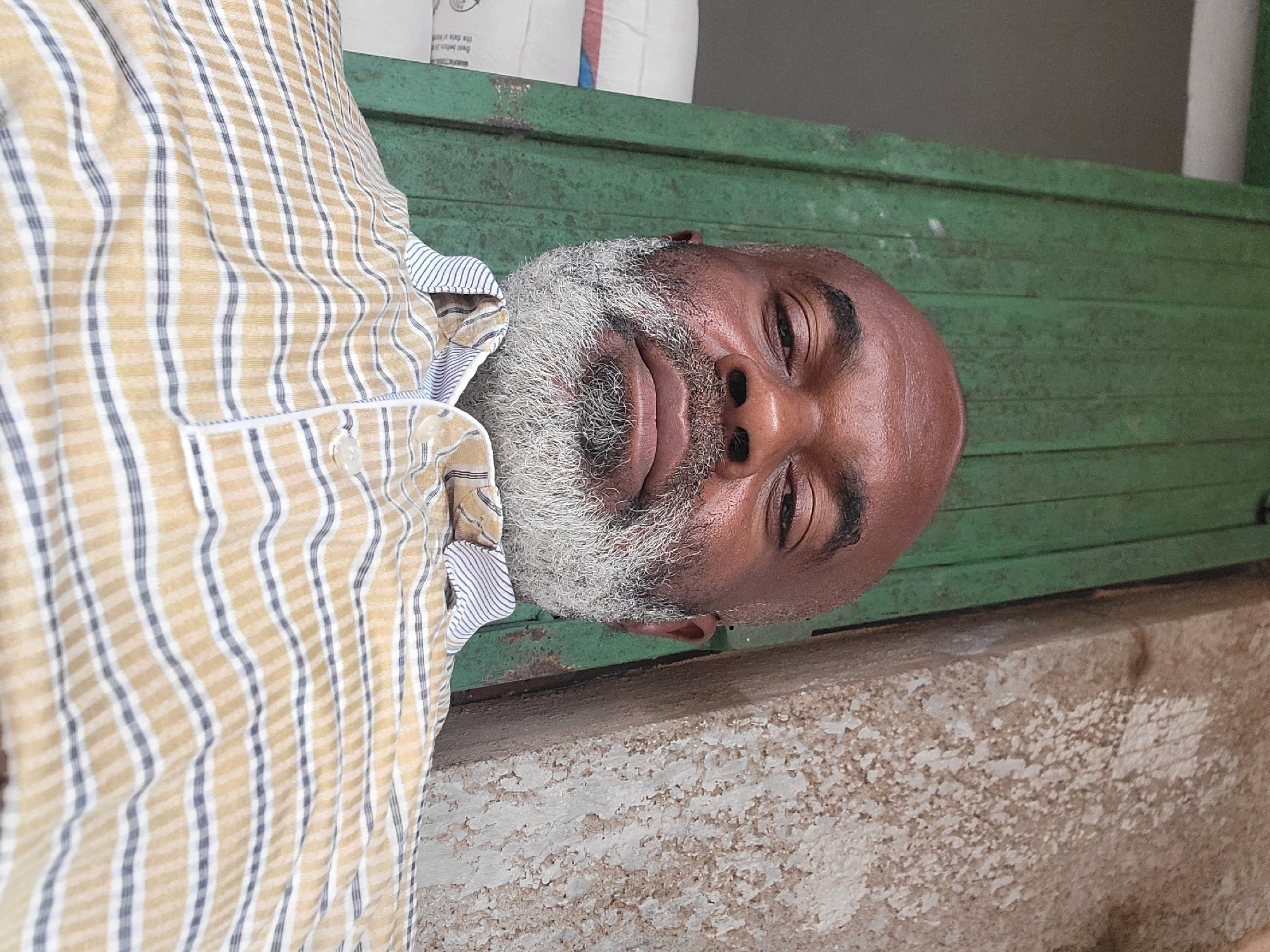 A close-up shot features a middle-aged Black man with a bald head and a white-gray beard. He's wearing a striped shirt with yellow and white tones. The background includes a weathered green wooden surface and a deteriorated concrete ledge. Based on the request for the location being Maiburiji, Nigeria, this scene is likely somewhere in the vicinity. No specific actions or interactions are discernible.