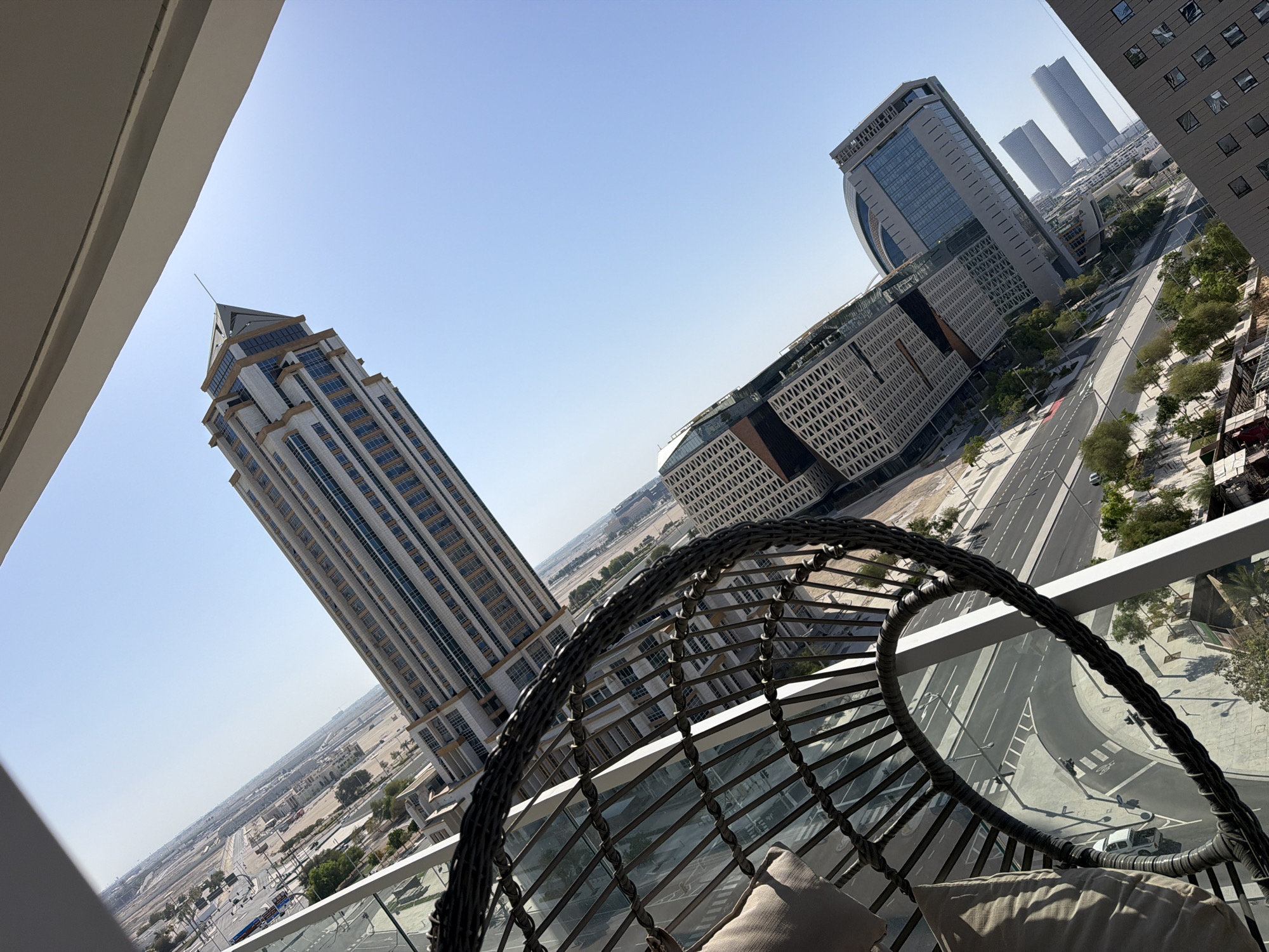 This image, taken from a high vantage point, showcases a modern cityscape under a clear blue sky. In the foreground, a portion of a woven chair and a cushion are visible, suggesting a balcony or outdoor seating area. Dominating the mid-ground are several contemporary buildings with sleek, glass facades and geometric designs. One particularly tall skyscraper with a distinctive pointed top stands out. Below the buildings, a road network with visible lanes and streetlights indicates urban infrastructure. The background reveals a sprawling, partially developed urban landscape with more buildings and arid terrain stretching towards the horizon. The overall impression is one of a prosperous, modern city with a clear, sunny day.