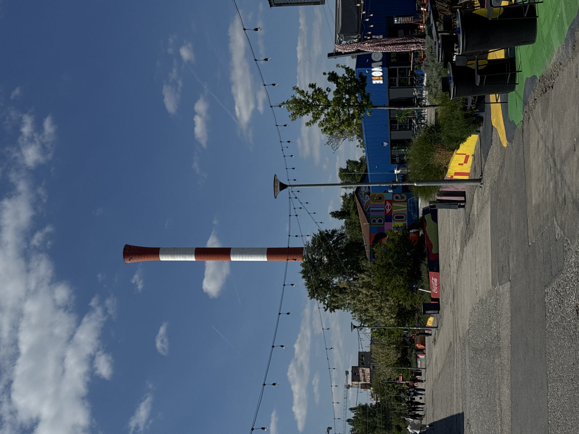 The image depicts an outdoor scene in Belgrade, Serbia, likely within the industrial-chic area of Savamala, known for its repurposed warehouses and vibrant cultural life.
The sky is a clear blue with scattered white clouds. A tall, industrial chimney painted with alternating bands of red and white stands prominently against the sky. Strung across the street are strands of decorative lights with small bulbs. A street runs along the right side of the image, with a variety of businesses and attractions lining it. These include structures made from shipping containers, painted murals, and signage like a "Big Love" sign and a Coca-Cola ad. Trees and other greenery are interspersed along the street. The street is made of concrete, with various tones. People can be seen walking in the distance.