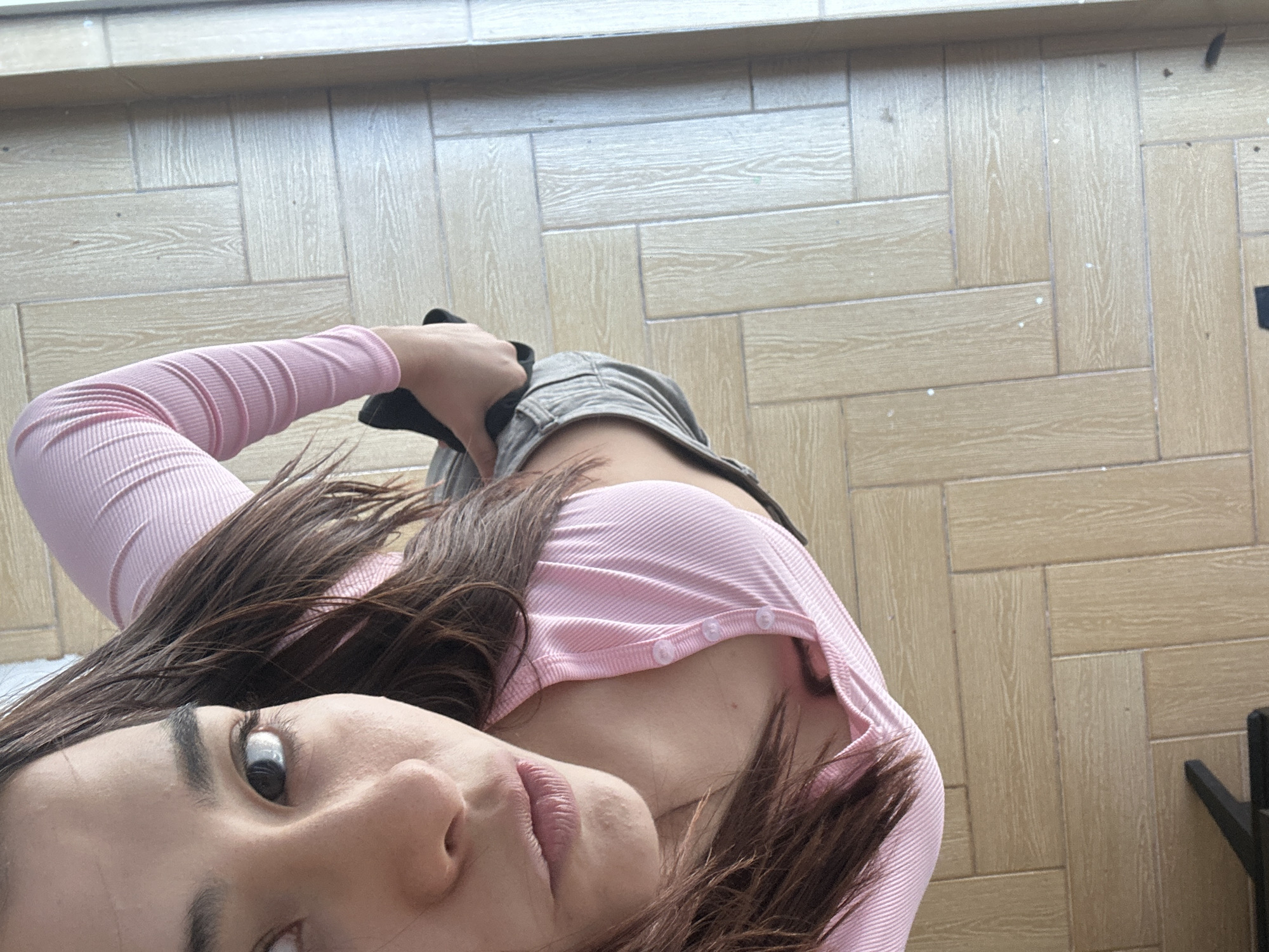 A selfie of a woman is taken from a high-angle perspective. The woman has fair skin, brown hair, and is wearing a pink long-sleeved shirt with buttons. She is wearing light gray denim shorts. Her face is positioned at the bottom left of the frame, with her gaze directed towards the camera. The background consists of a wooden-patterned floor. The location is unknown.