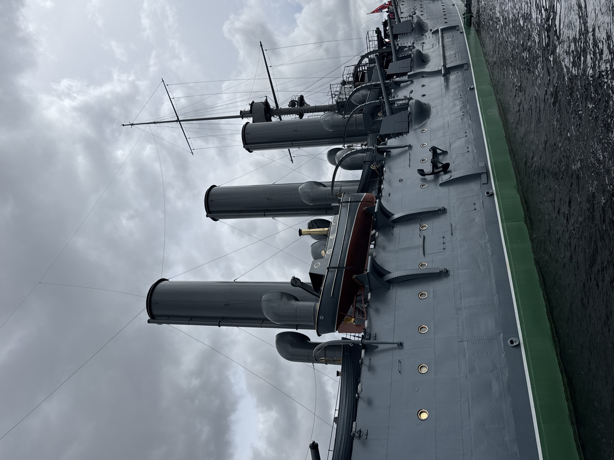 The image shows the Russian protected cruiser Aurora, a museum ship, moored on the Neva River in Saint Petersburg, Russia. The angle of the photograph is vertical, looking along the port side of the vessel from bow to stern. Four prominent, grey smokestacks are visible, ascending from the deck towards a cloudy, overcast sky. A lifeboat is positioned between the third and fourth smokestacks. The vessel's gray hull is lined with rivets and circular ports, some of which appear to be illuminated. Black anchors are visible along the hull. The vessel is bordered by the dark water of the Neva River.