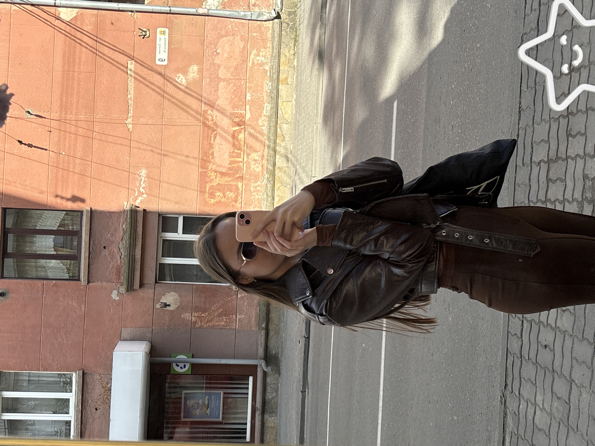 A young woman with long brown hair, wearing a brown leather jacket and brown pants, takes a mirror selfie with her pink phone in front of a pink building. She is standing on a sidewalk with a street in the background. The building appears to be in Ternopil, Ukraine, as evidenced by the Ukrainian street sign on the building.