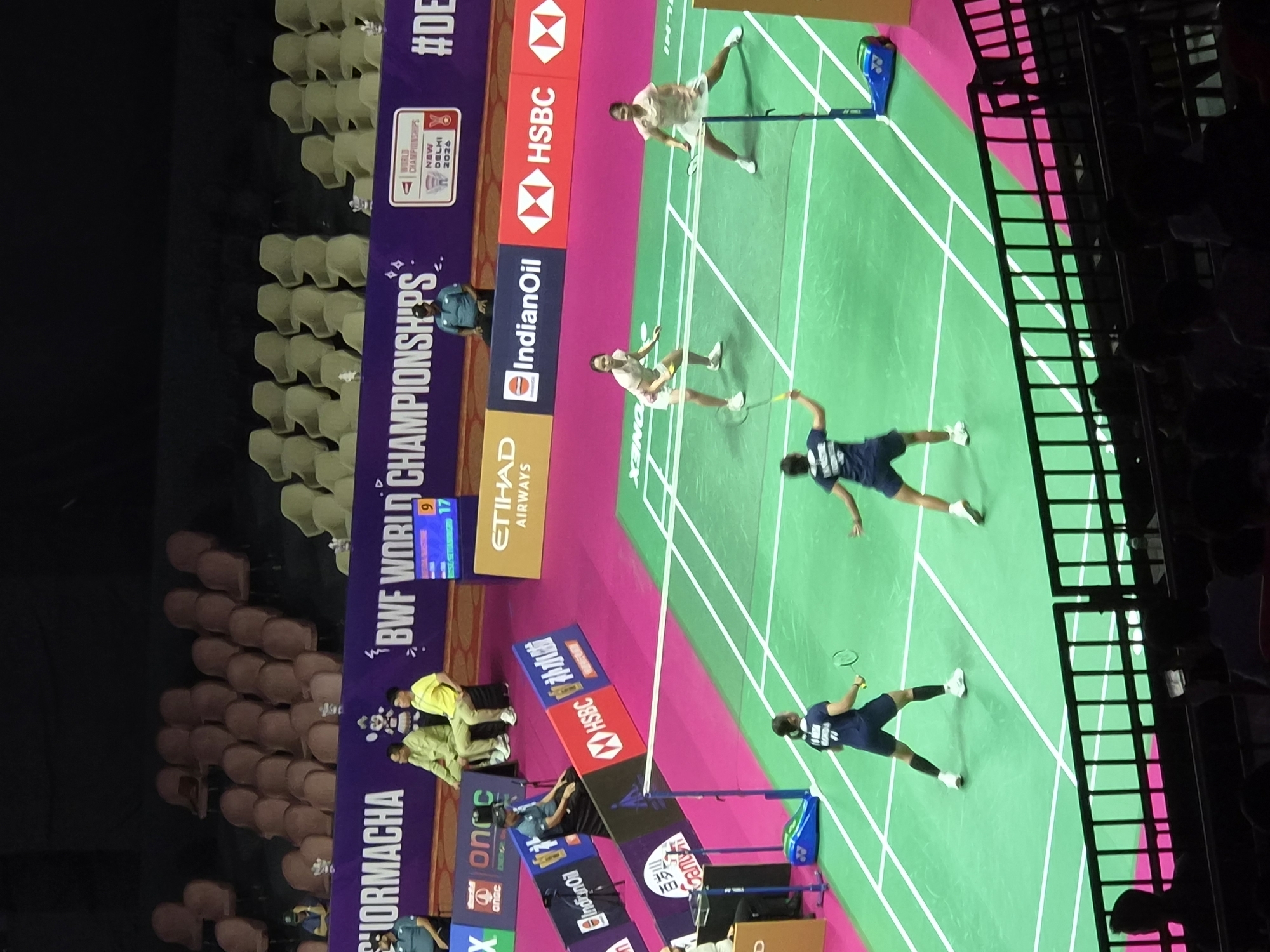 The image depicts an indoor badminton match at the Indira Gandhi Indoor Stadium in Delhi, India. The court is green with white lines, and the net is visible. Four female athletes are on the court, two on each side, engaged in the game. Two players in dark blue uniforms are on one side, and two players in light-colored uniforms are on the other. One player in a dark blue uniform is in motion, preparing to hit a shuttlecock, while another player in the same uniform is in a defensive stance. The players in light-colored uniforms are also actively participating in the rally. Spectator seating is visible in the background, with empty and occupied seats. Advertising banners are displayed around the court, including "BWF WORLD CHAMPIONSHIPS NEW DELHI 2026," "ETIHAD AIRWAYS," and "IndianOil." The lighting is typical of an indoor sports arena.