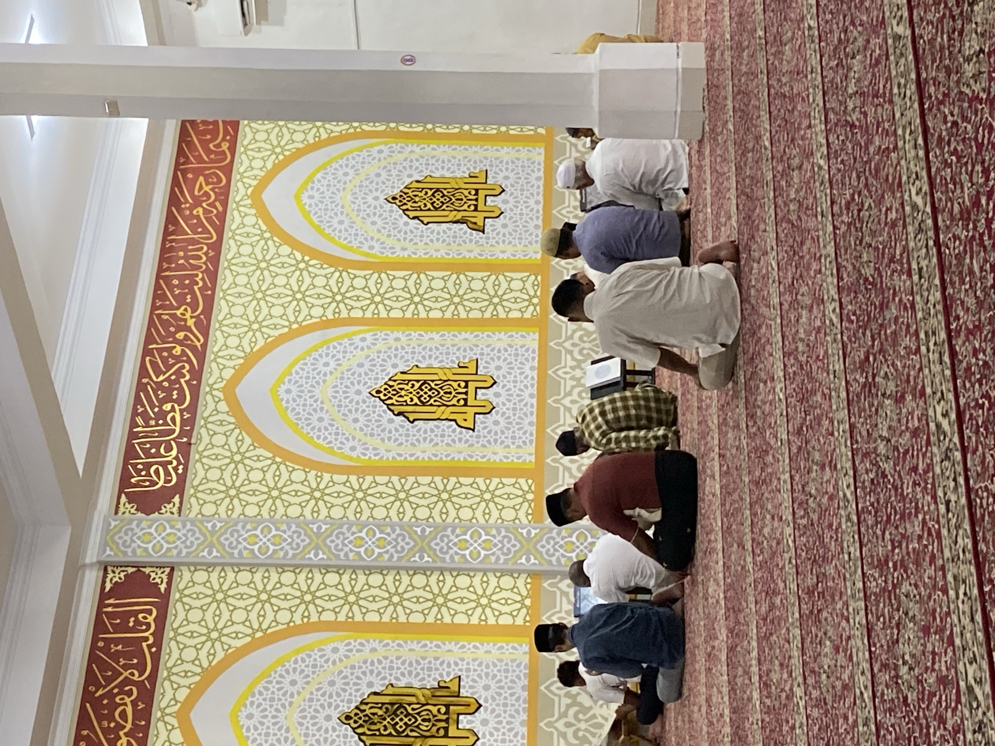 The image depicts the interior of a mosque with several men seated on a red carpet, facing a wall adorned with Islamic calligraphy and geometric patterns. The wall features three arched decorative niches, each containing a stylized golden emblem. The men are in various stages of prayer, with some in a seated position and others leaning forward. The environment appears to be a place of worship. While the image itself does not provide specific location markers, it is described as being in the city of Lhokseumawe, Indonesia, suggesting this is a mosque within that city.