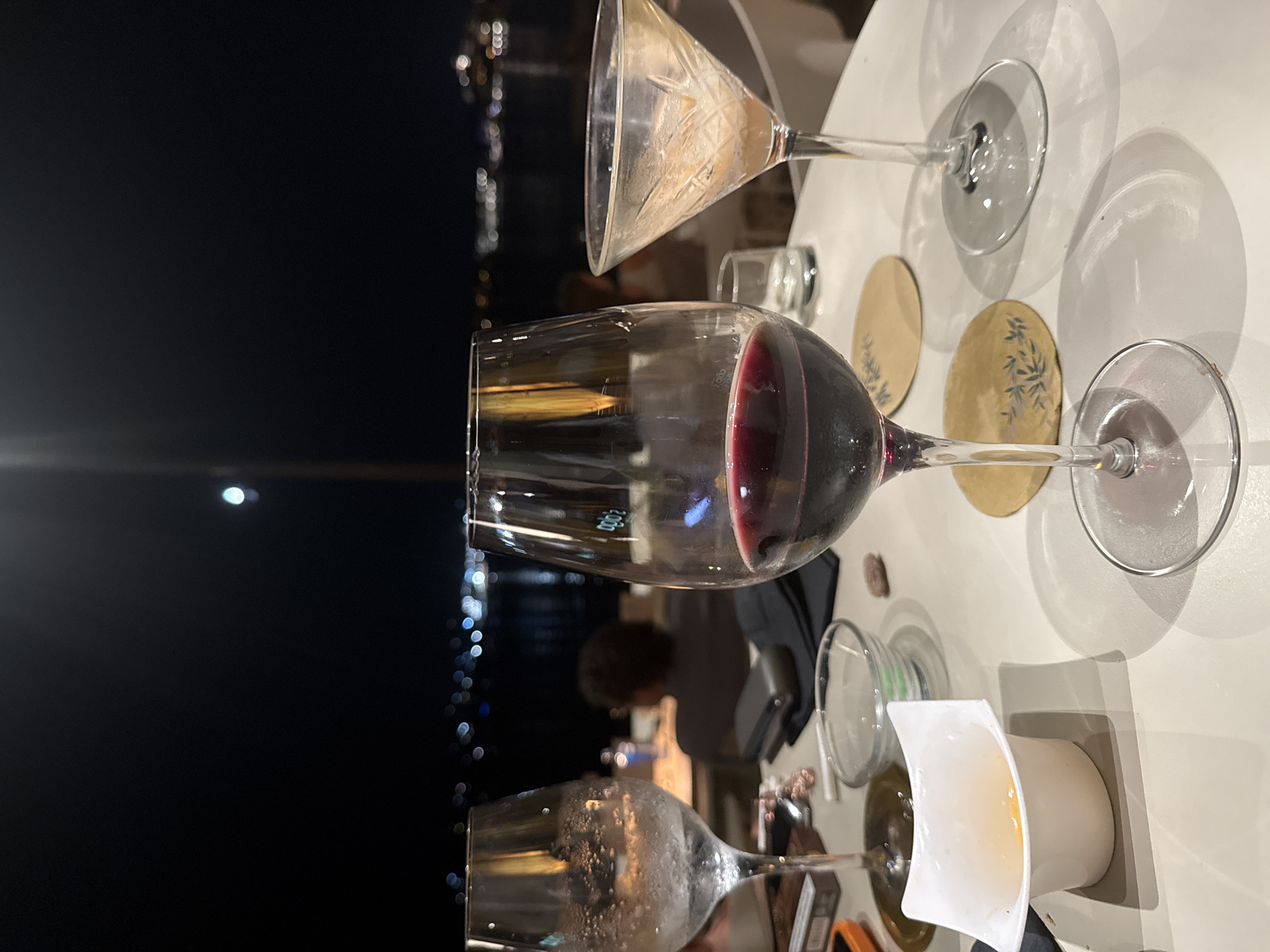 This image depicts an outdoor table setting at night, likely in Eivissa, Spain, with a blurred cityscape and lights visible in the background. The table holds three glasses: one filled with red wine, another appearing to be an empty martini glass with residue, and a third frosted glass with water or ice. Small dishes, coasters, and what appears to be a cell phone are also present on the table. A person is partially visible in the background, seated at another table. The lighting suggests a restaurant or bar environment.