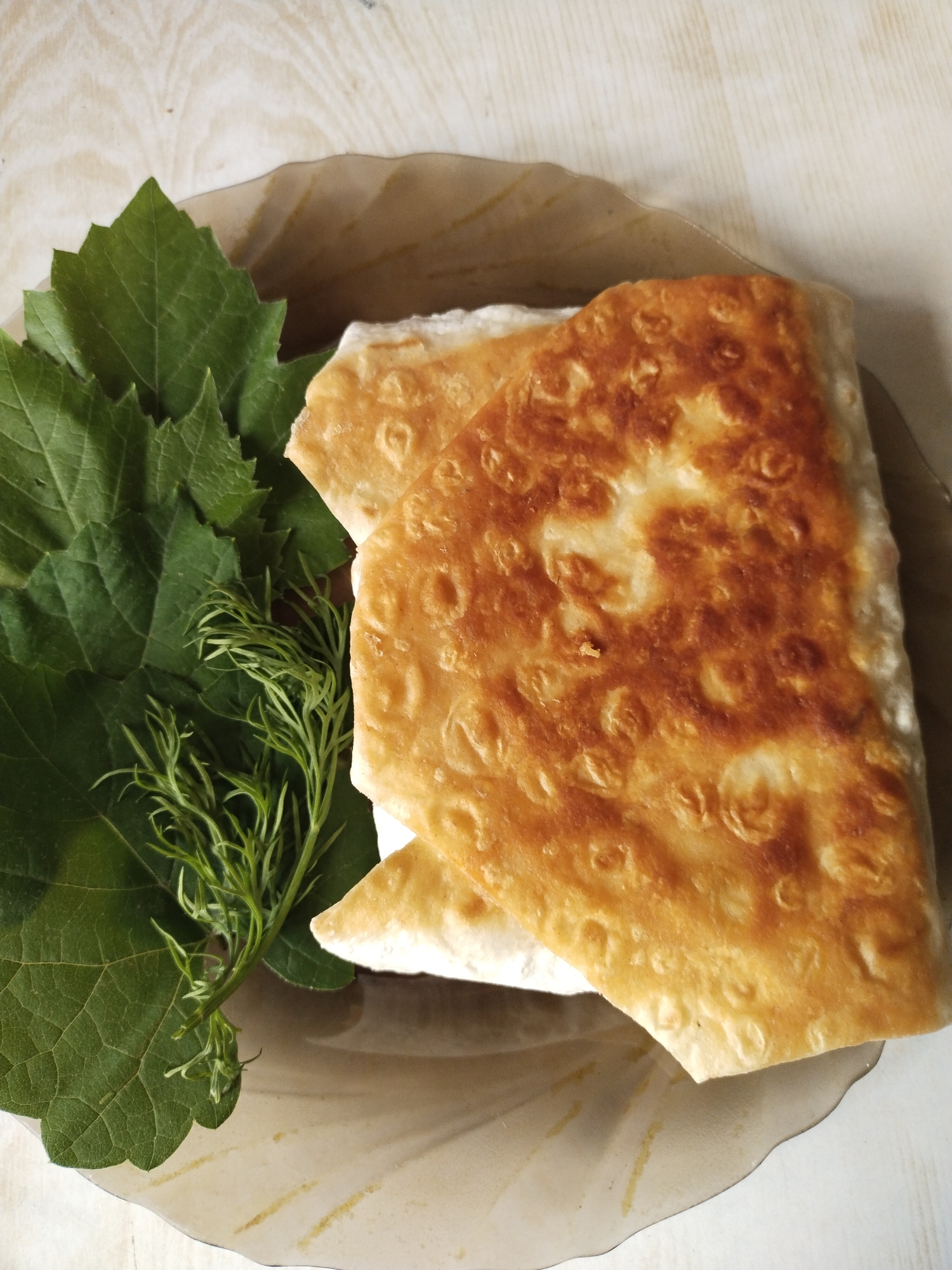 An overhead shot displays a dish consisting of two pieces of golden-brown, folded pastry, likely a type of lavash or flatbread, arranged on a translucent, light-brownish fluted plate. The pastry appears freshly cooked, with a crispy, somewhat bubbly texture and varying shades of golden-brown from frying or baking. A white filling is visible peeking out from the edges of the pastry, suggesting cheese or a similar dairy product.
Accompanying the pastry on the left side of the plate are fresh green garnishes: several broad, dark green leaves, which resemble grape leaves, and a sprig of lighter green, feathery dill. The greens appear vibrant and fresh.
The plate rests on a light-colored surface, possibly wood or laminate, which has a subtle linear grain texture. The overall presentation suggests a prepared meal, possibly a breakfast, lunch, or snack. No specific activity is depicted other than the food being served. There is no visible text in the image. The food, particularly the combination of lavash-like pastry, fresh greens, and dill, is consistent with culinary traditions found in Eastern Europe or the Caucasus region, aligning with the provided location context of Horishni Plavni, Ukraine.