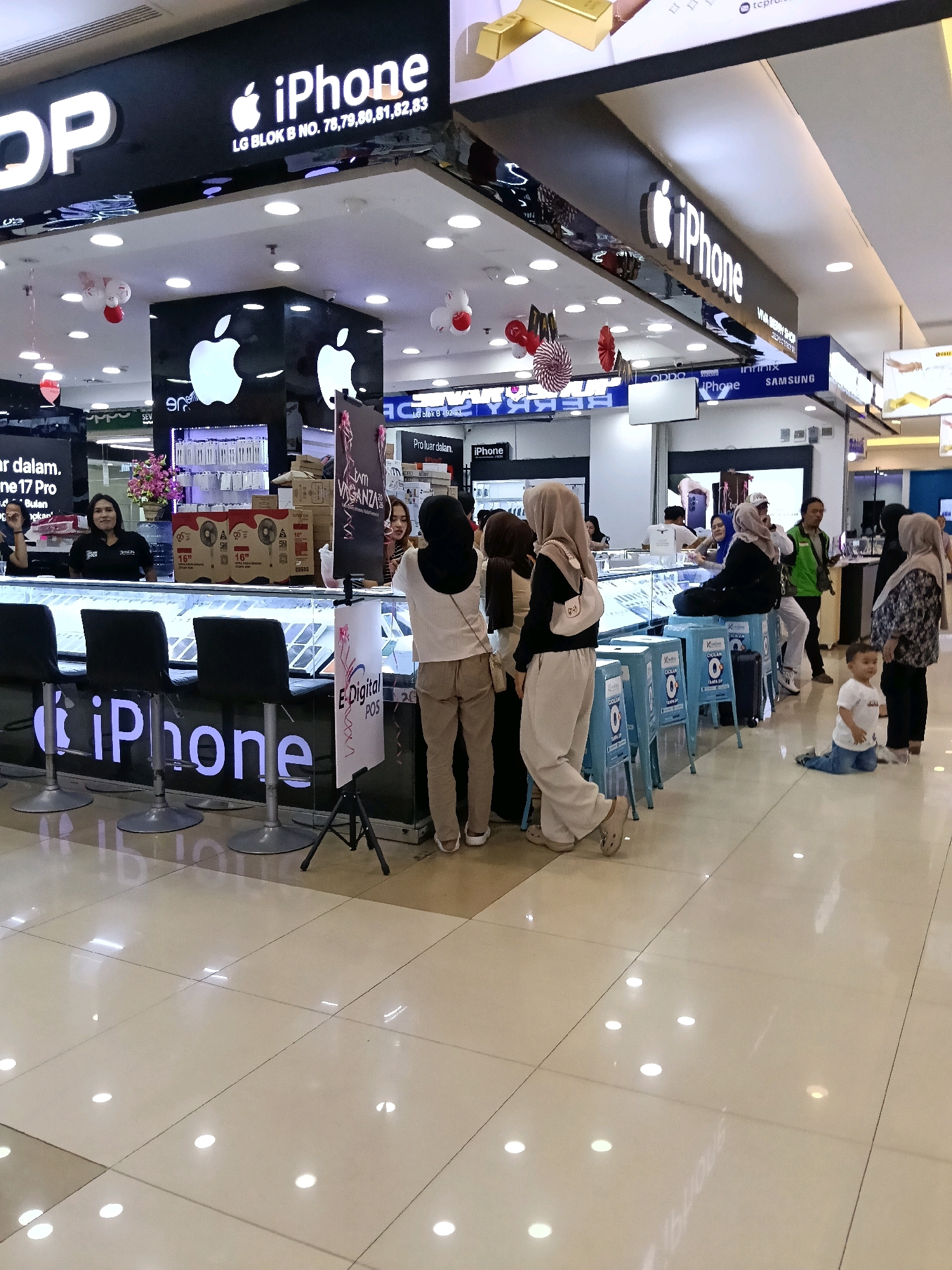 The image depicts the interior of an electronics store, likely located in a mall, in Tangerang, Indonesia, specifically within the Supermal Karawaci. The store prominently features an "iPhone" branded counter with multiple display cases showcasing electronic devices. Several individuals are present: two female employees are behind the counter, interacting with customers. A group of customers, including women and a child, are seen browsing or making purchases. The environment is brightly lit with fluorescent lighting on the ceiling, and the floor is a polished tiled surface. Signage for various brands like "OPPO," "SAMSUNG," and "iPhone" is visible. The overall scene suggests a commercial setting focused on the sale of mobile phones and related accessories.