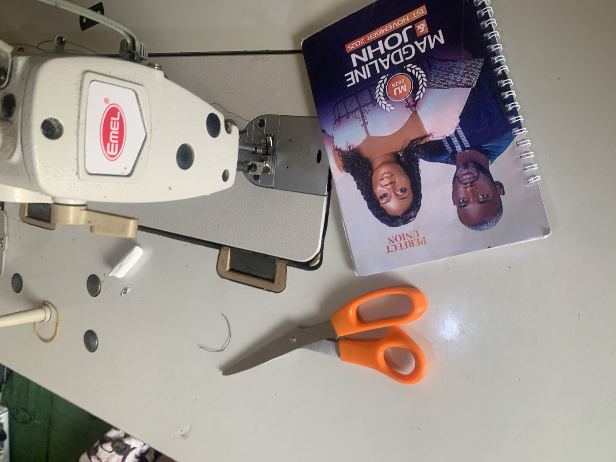 In Jauro Yinu, Nigeria, a sewing machine is positioned on a white surface. Next to it, a pair of orange-handled scissors rests on the surface, along with a small piece of white chalk and a strand of dark thread. To the right, a notebook is placed on the surface, open to a page displaying a couple's photograph. The notebook has text that reads "PERFECT UNION" and "MAGDALINE & JOHN 1ST NOVEMBER 2025". The cover of the notebook is dark blue and features the initials "MJ" within a circular emblem, along with the year "2025".