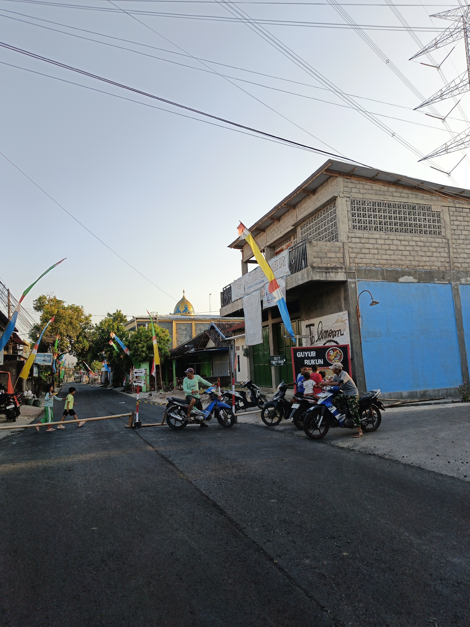 A street in Trucuk, Indonesia is depicted with several motorcycles and people. A temporary road closure, made of bamboo poles and red and white markers, is set up across the asphalt road. Several colorful flags are erected along the sides of the street. In the background, a building with a blue wall and a brick upper story stands. A mosque with a golden dome is visible further down the street. Several people are gathered around motorcycles, and two children are walking across the road. The sky is clear and bright, suggesting it is daytime. The image appears to be taken from a slightly low angle.