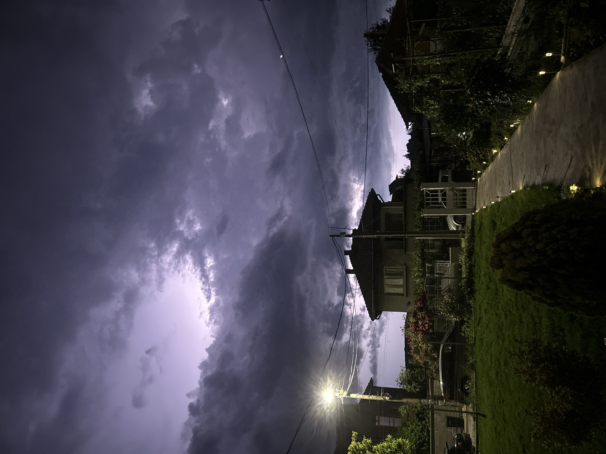 The image shows a nighttime scene in what appears to be a residential area, possibly in Burgas, Bulgaria, given the prompt. Dark, ominous clouds dominate the sky, with a visible flash of lightning illuminating them from within. The image is taken from a low angle, looking up at buildings and utility lines. A streetlight shines brightly, casting a glare and illuminating nearby trees and structures. To the right, a path or walkway is lined with small, evenly spaced lights. Vegetation, including trees and bushes, is visible on both sides of the path. The houses have multiple windows. A vehicle or other large object is partially visible behind the vegetation.