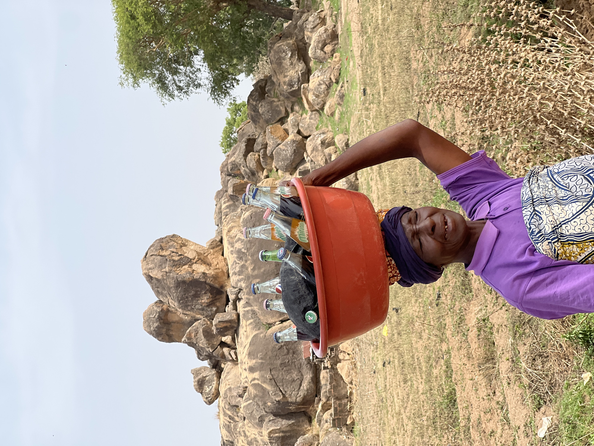 A woman is pictured outdoors, carrying a red tub full of glass bottles on her head. She is smiling, with her right arm raised. She is wearing a purple shirt, a dark-colored head wrap, and a patterned cloth around her waist. The background includes rocky terrain and a tree. The photograph appears to be taken in a rural setting, possibly in or near Udu, Nigeria. The foreground consists of dry grass and dirt.