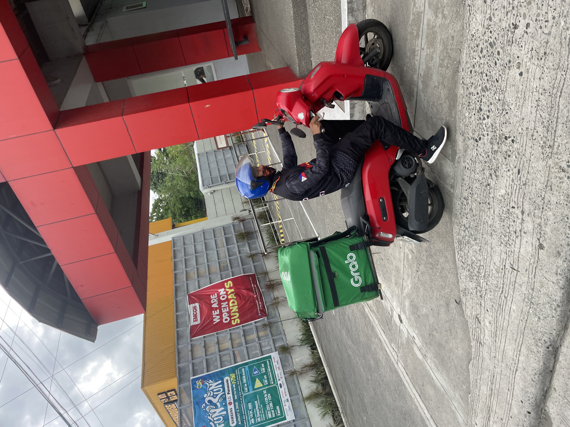 A Grab delivery rider wearing a blue helmet and a dark long-sleeved shirt with a circular logo on the left sleeve and the Philippine flag on the right sleeve, is riding a red scooter with a large green Grab food delivery bag attached to the back. The rider is wearing black pants and white sneakers with a black logo. The scooter has a black seat and a license plate. The rider is on a concrete road in front of a building with a red and yellow exterior. A red banner on the wall says "EMCOR WE ARE OPEN ON SUNDAYS". Two other posters are visible on the wall. The sky is overcast. The scene appears to be in Davao City, Philippines, given the presence of Grab, a Philippine company, and the overall architecture of the buildings.