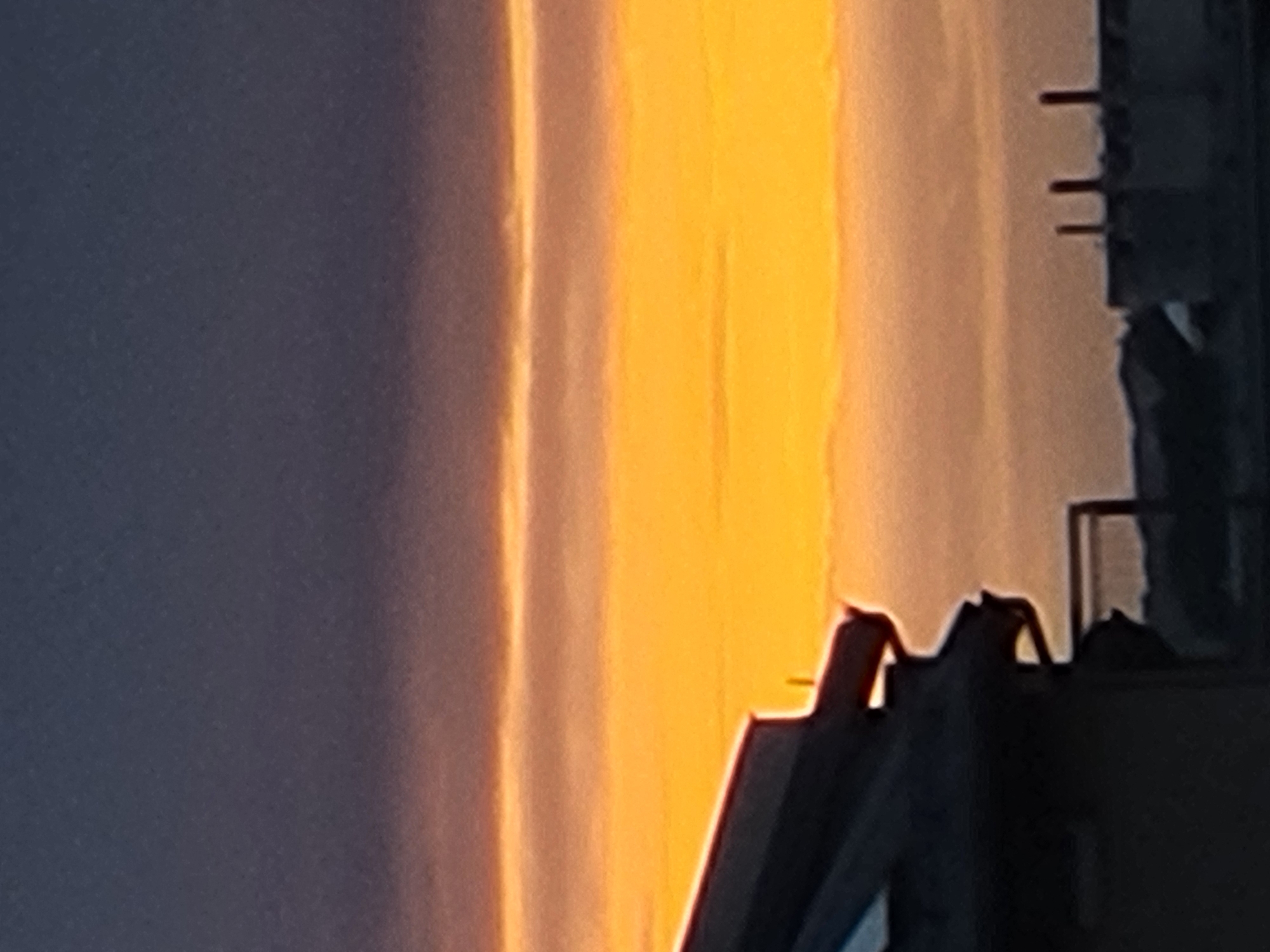 A silhouetted image of rooftops and industrial structures against a vibrant sunset sky. The sky transitions from a deep, dusky blue at the top to a bright orange and yellow band, with thinner layers of muted orange and purple clouds in between. In the foreground, the dark silhouette of a building with a sloped roof and an overhang is visible on the left. To the right, the outlines of several industrial chimneys or stacks rise against the colorful sky, suggesting an urban or industrial setting. The overall impression is one of a twilight cityscape.