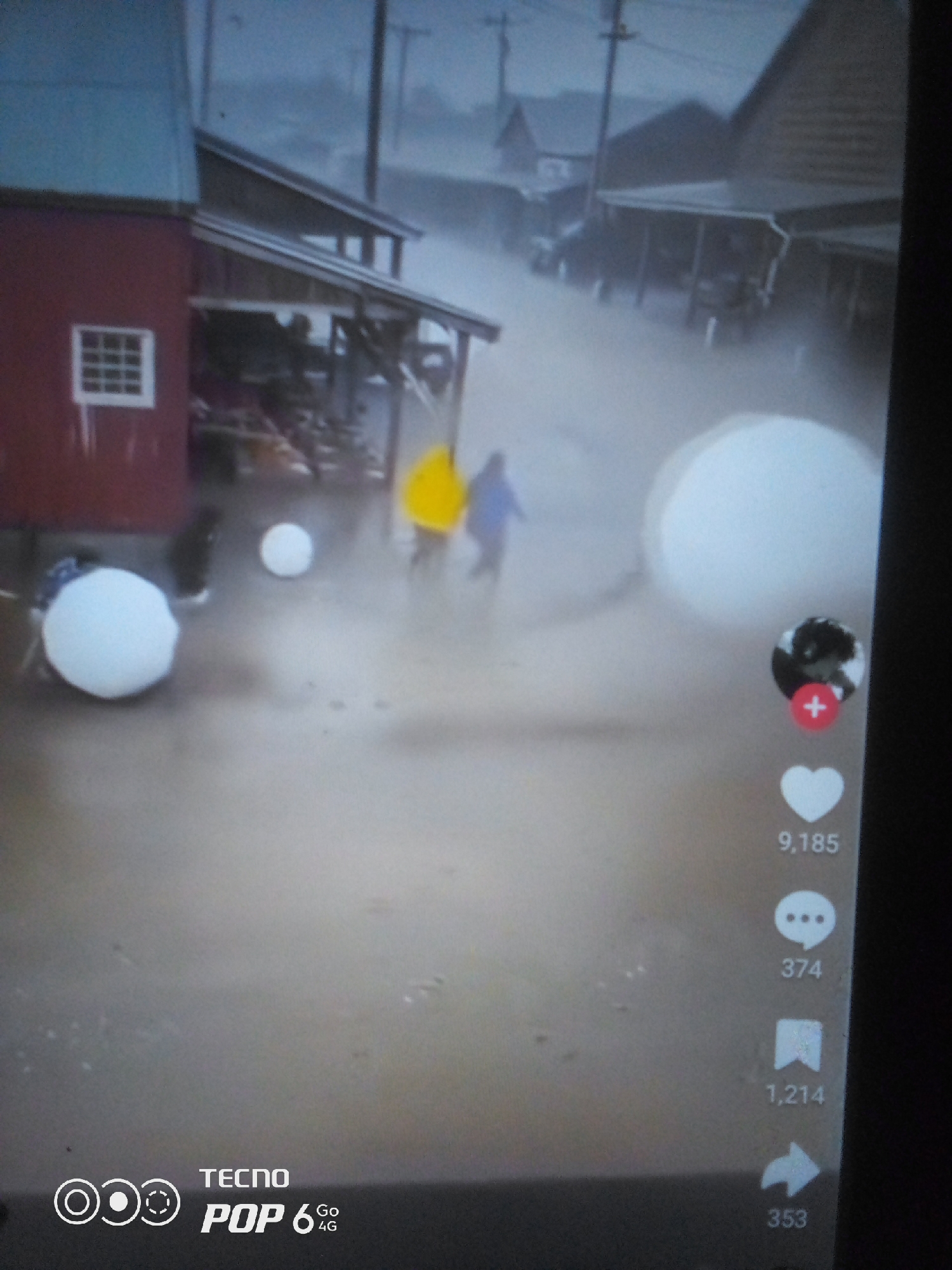 This is a forensic description of an image that appears to be a still frame from a video, likely from a social media platform, showing a flooded street. The scene is set in what is identified as Howayi, Nigeria, during what seems to be a downpour or heavy rain, as indicated by the blurry, misty atmosphere and the water covering the ground.
In the foreground, on the left side of the image, is a red building with a white-gridded window. Adjacent to it, a covered walkway or shed extends outwards, supported by posts. The ground is inundated with water, reflecting the dimly lit environment. Two large white spherical objects are visible in the water near the red building. Further into the scene, two individuals are walking through the floodwaters. One person is wearing a bright yellow raincoat, and the other is in a blue jacket. They appear to be moving away from the camera.
In the background, on the right side, are more structures, possibly shops or houses, with similar covered walkways. The environment is characterized by low visibility due to the rain and fog.
On the right edge of the frame, social media interaction icons are superimposed on the image. These include a profile picture with a '+' icon, a heart icon with the number "9,185" below it, a speech bubble icon with "374" below it, a bookmark icon with "1,214" below it, and a share arrow icon with "353" below it.
At the bottom left corner of the image, a logo is visible, featuring concentric circles and the text "TECNO POP 6 Go 4G". This logo, along with the social media icons, suggests the image was captured or shared via a Tecno mobile device on a social media platform. The overall impression is one of a localized flooding event in an urban or semi-urban area in Nigeria.
