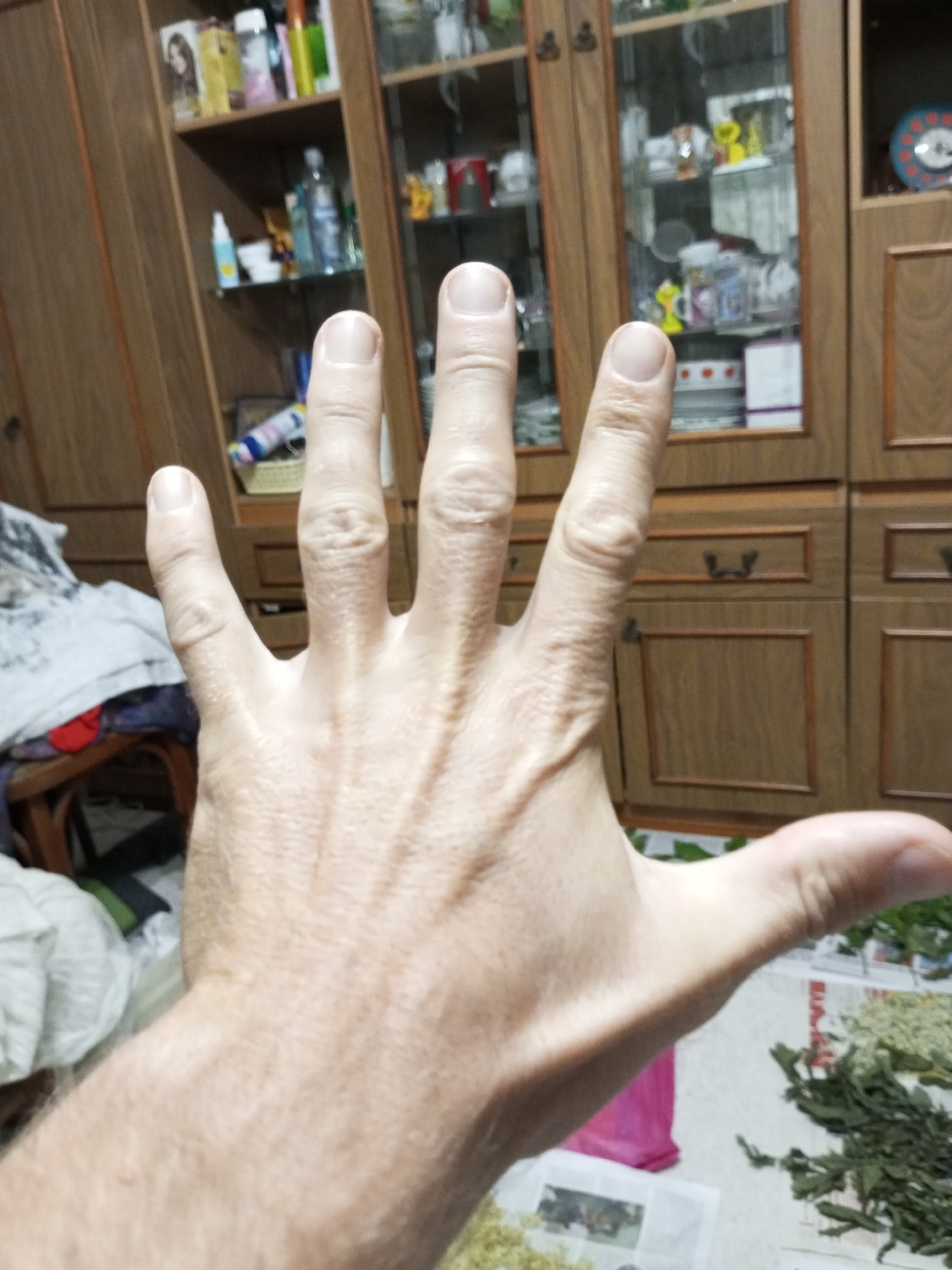 A close-up, eye-level shot shows the back of a person's left hand with fingers spread, palm facing the camera. The hand is positioned in the foreground, obscuring the background, which appears to be a room with wooden furniture and shelves filled with various items. The overall lighting is dim. The image does not provide enough information to determine the location within Ароматновское сельское поселение, Ukraine.
