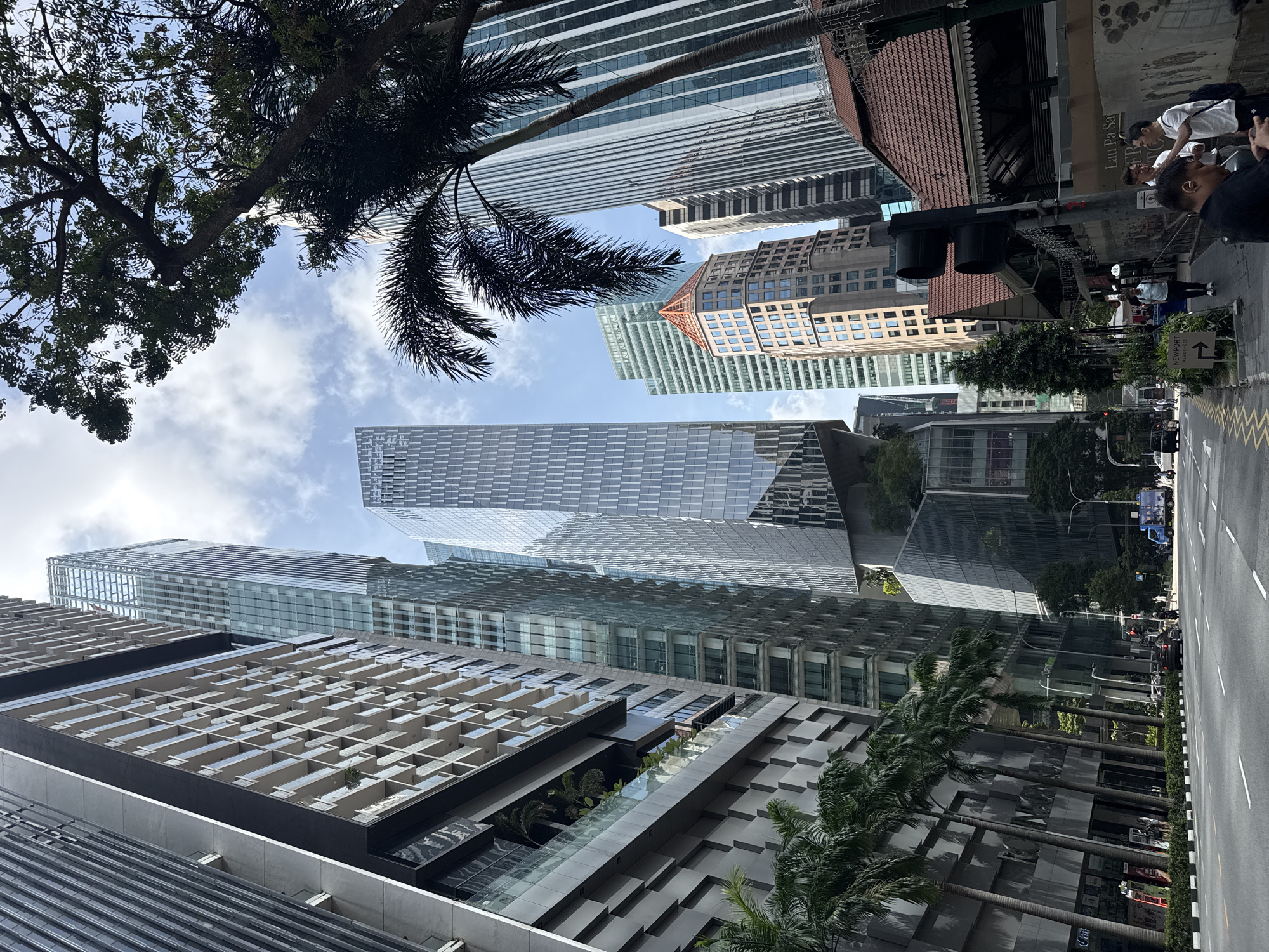 This is a low-angle, eye-level shot of a street in Singapore, Singapore, with tall modern skyscrapers dominating the skyline. The buildings are made of glass and steel, with intricate architectural designs. Palm trees line the street, adding a tropical touch to the urban environment. In the foreground, a road with cars and pedestrians is visible. The sky is partly cloudy, with the sun casting shadows on the buildings. A traffic light is also visible on the right side of the image. The overall scene depicts a bustling city with a blend of modern architecture and natural elements.