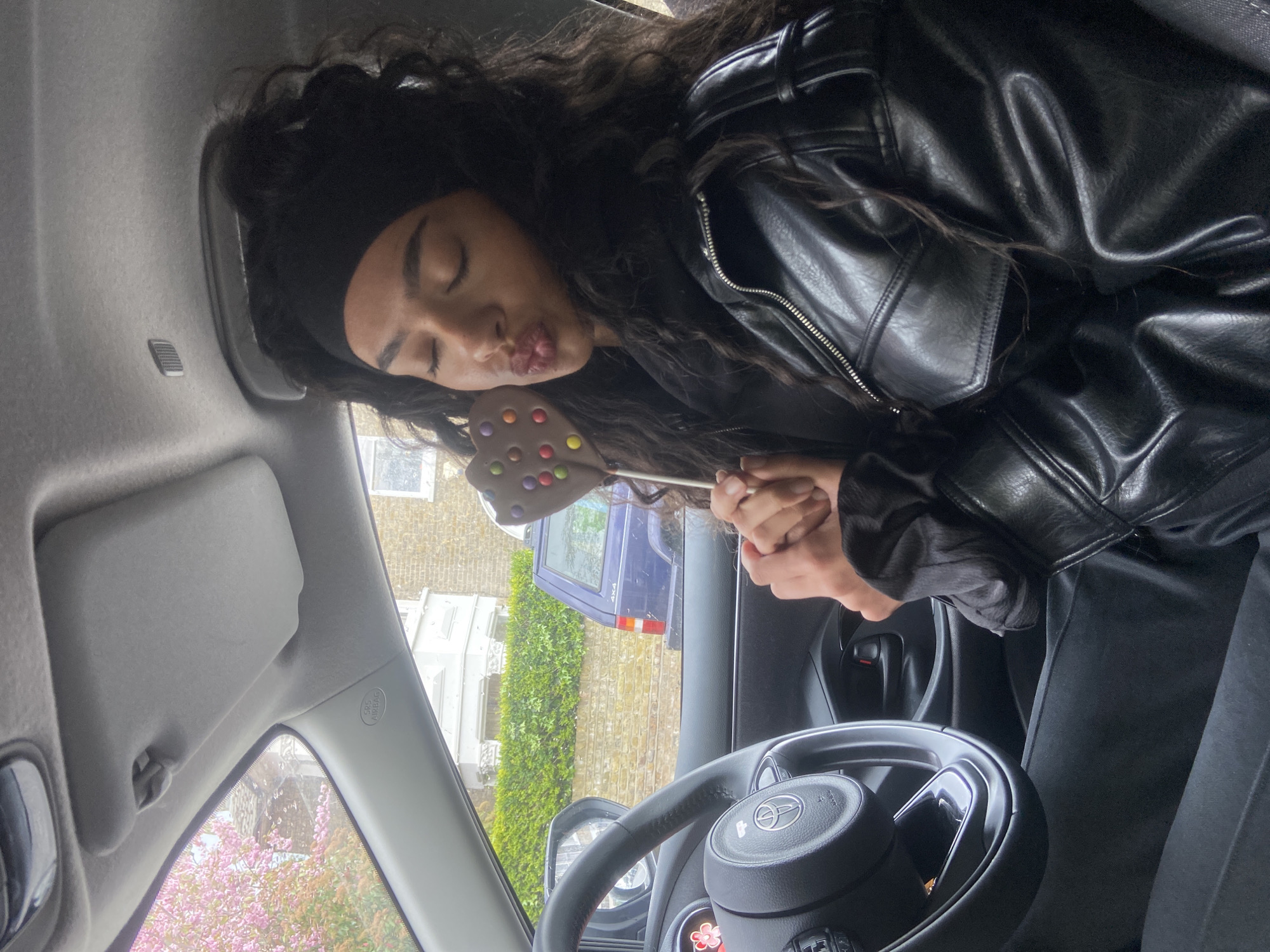 A young woman with dark curly hair and a black headband is sitting in the driver's seat of a car. She is wearing a black leather jacket and holding a heart-shaped chocolate lollipop with colorful candy dots. She is making a kissing face towards the lollipop. The car's steering wheel, dashboard, and sun visor are visible. Outside the car window, there is a view of a street with brick buildings, greenery, and a parked blue SUV. The location is suggested to be in London, United Kingdom, based on architectural cues common to the area.