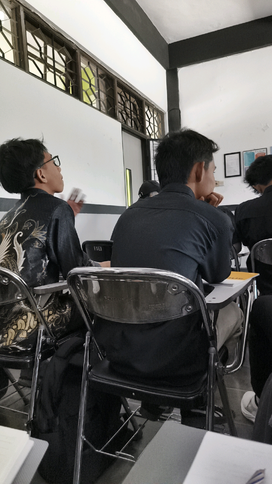 The image depicts an indoor scene, likely a classroom or meeting room, in Kartasura, Indonesia. In the foreground, two individuals are seated at desks, viewed from the back. The person on the left is wearing glasses and a black patterned shirt, looking to their right while holding a white object. The person on the right is wearing a black collared shirt and has their hand resting on their chin, looking forward. Their chairs appear to be black metal frame with black seats and backs, with "FORTUNER" and manufacturing details visible on the chair back on the right. In the background, other individuals are also seated at desks. A black and white striped beam runs across the ceiling. To the left, a row of windows with a geometric lattice pattern is visible. The walls are white. Small framed pictures are mounted on the wall in the upper right corner.