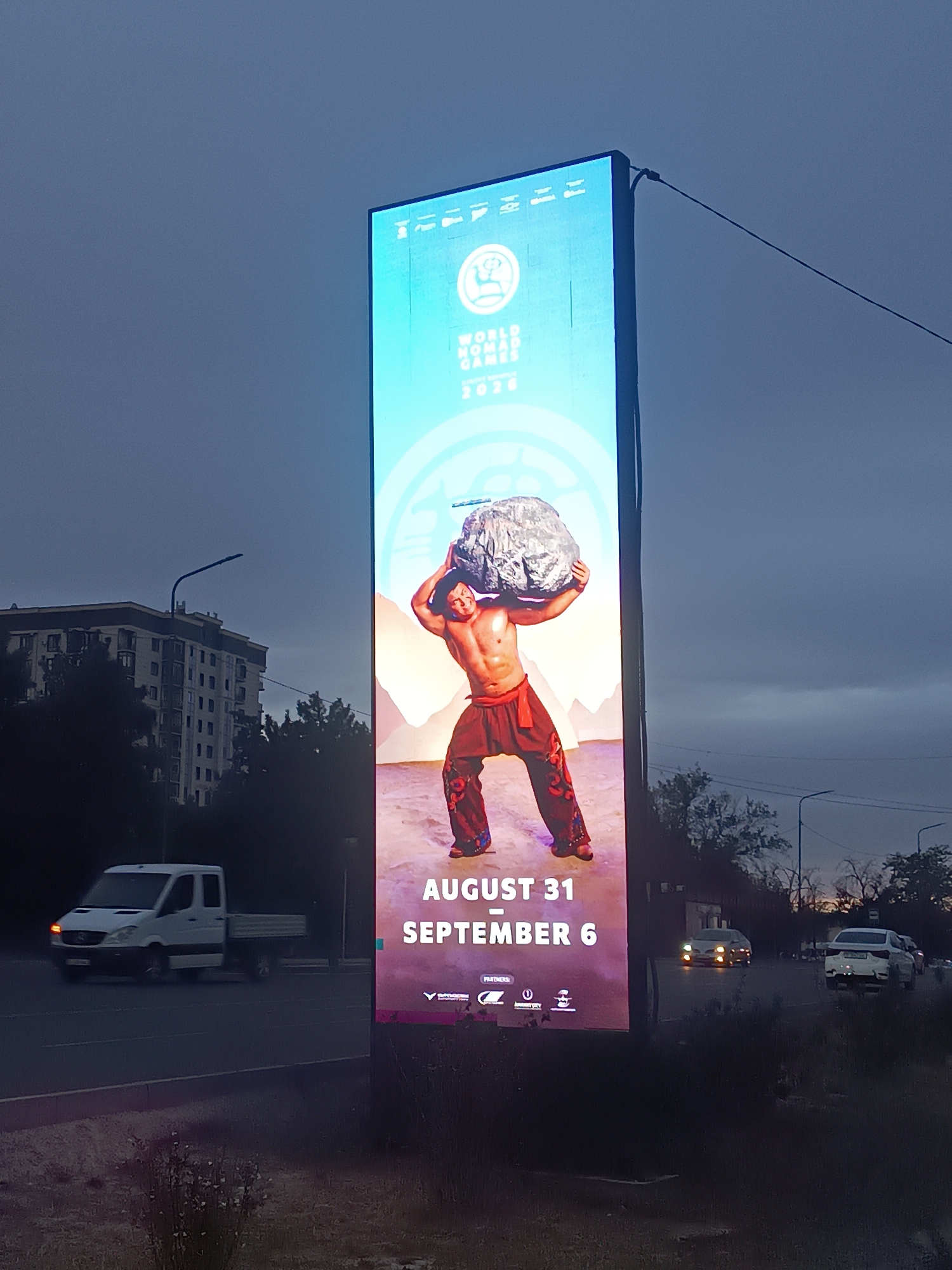 An outdoor digital billboard displays an advertisement for the World Nomad Games 2026. The billboard features a muscular man with his torso bare, wearing ornate trousers, lifting a large, rough stone above his head. Text on the billboard reads "WORLD NOMAD GAMES 2026" and "AUGUST 31 SEPTEMBER 6". Below the dates are logos for partners, including one with the word "PARTNERS". The billboard is situated on a city street in Bishkek, Kyrgyzstan, evidenced by the presence of apartment buildings, streetlights, and passing vehicles. A white van is visible on the left side of the road, and several cars are driving on the road in the background. The sky is overcast and appears to be dusk or dawn. The overall scene is a typical urban streetscape with a prominent advertisement.