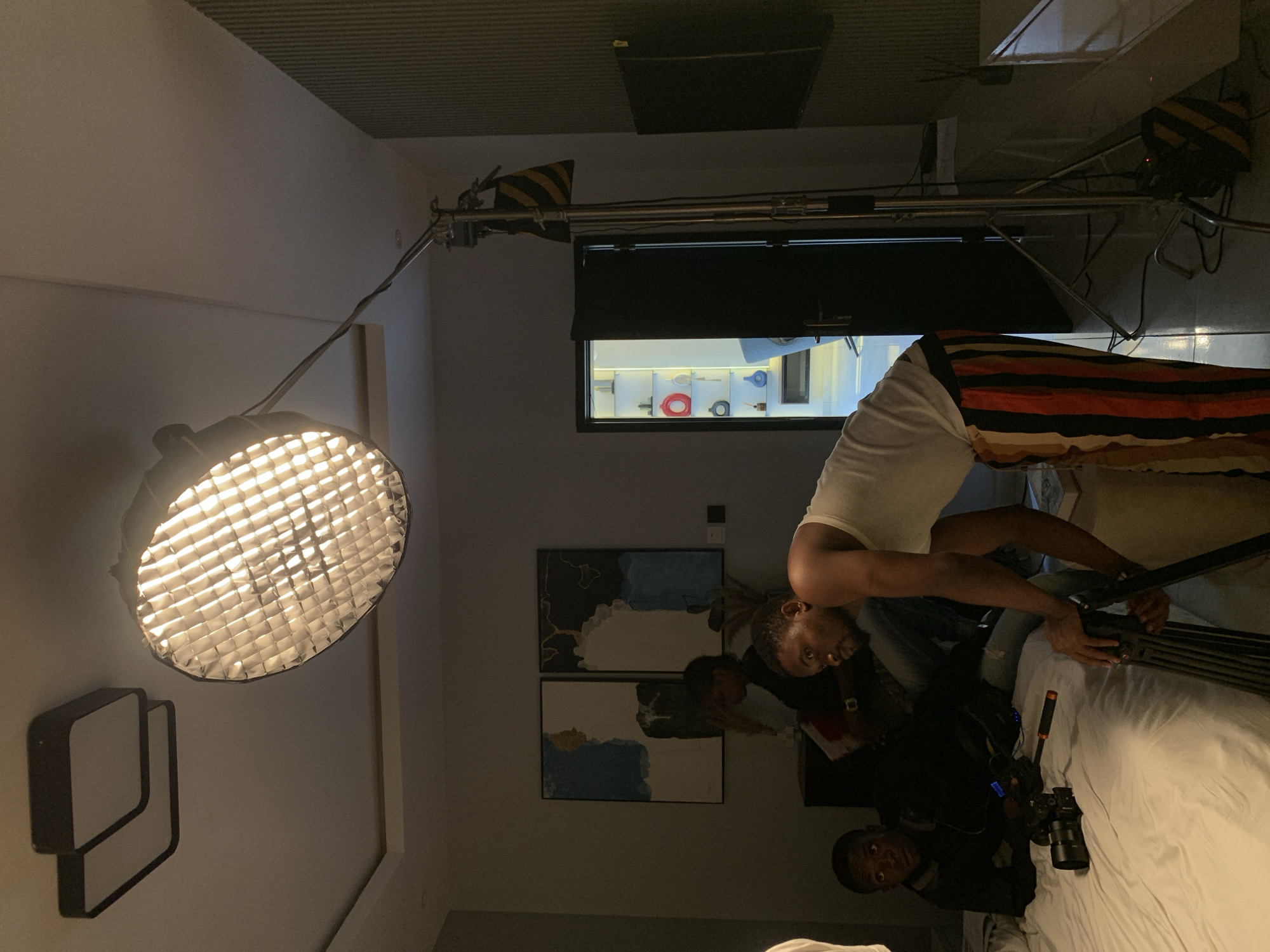 This image depicts an indoor scene, implicitly located in Moba, Nigeria, resembling a film or photography production setup within a residential or studio environment. A prominent, large, circular studio light with a honeycomb grid is actively illuminated on a C-stand, positioned centrally to the right. In the foreground, a male subject with dark skin and a beard, wearing a white tank top and multi-colored striped pants (red, orange, black, beige), is leaning forward, attending to a black tripod. To his left, another male subject, also with dark skin, wearing a dark long-sleeved shirt and blue jeans, is seated on a bed covered with white bedding, looking directly forward. A camera with a lens attachment is visible on the bed near this individual. A third person is partially visible, seated in the background. The room features dark gray walls; one wall displays two abstract art pieces, while an adjacent wall exhibits vertical textured paneling. A dark, open doorway on the right reveals a brightly lit adjacent room with white shelves holding various decorative items, including red and blue vases. Overhead, the ceiling is white with recessed lighting and two square light fixtures. A wall-mounted, rectangular black object, possibly a television or monitor, is visible on the textured wall. The overall scene suggests a controlled lighting environment for a production.