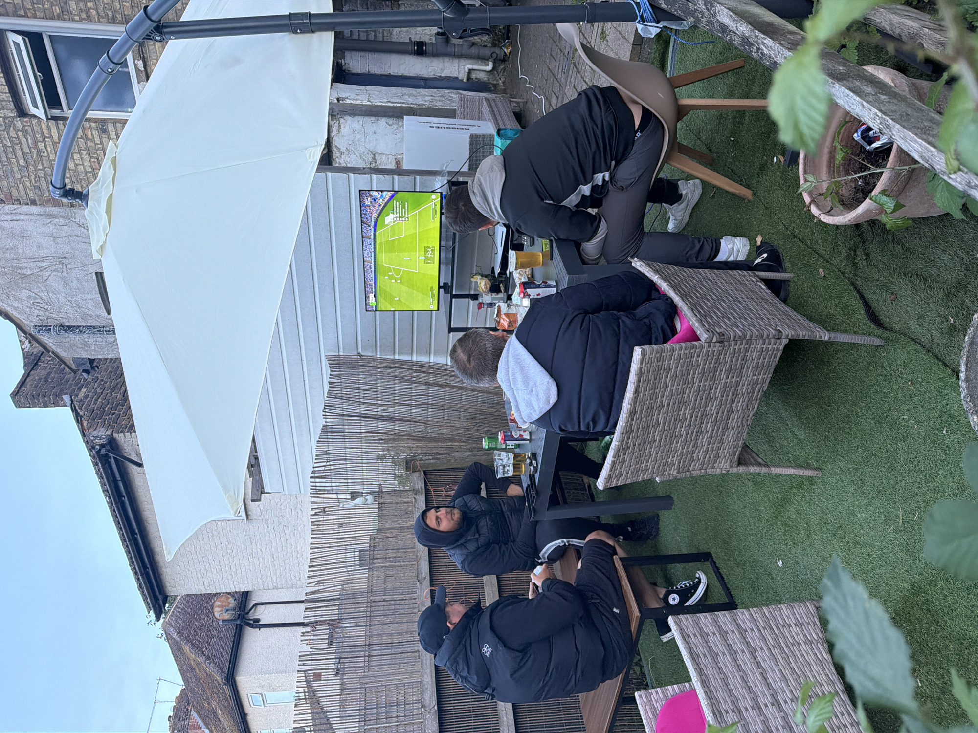 The image depicts an outdoor gathering of four adult males in a residential backyard, consistent with common architecture found in the United Kingdom. There is no visual information within the image to confirm the specific location as Maidstone. The scene is set on artificial green grass, with a large cream-colored cantilever patio umbrella providing shade, its dark grey pole visible on the right side. A black rectangular outdoor table serves as the central point for the group. Four men are present: 1. On the left, a man is seated on a wooden bench, facing partially away from the camera. He wears a black baseball cap, a black puffer vest over a black long-sleeved shirt, dark shorts, and black high-top Converse sneakers. He holds a small white object in his hands. 2. Seated at the table, facing slightly to the right, is a man with a beard, wearing a black hooded sweatshirt with the hood up, a black puffer vest, and black track pants featuring a white side stripe. 3. In the left foreground, partially obscured, a man is seated in a grey wicker-style chair at the table, holding a pink cushion or blanket. 4. On the right side of the table, a man is seated on a light brown plastic chair, with his back mostly to the camera. He is wearing a grey hooded sweatshirt over a black and white long-sleeved top and dark trousers, with his lower back partially exposed. All four men are directed towards a large flat-screen television, mounted on a black stand to the right of the table. The TV displays a football match, appearing to be a video game due to the on-screen graphics, including a score of "2-0" and "84'" (84th minute), along with clear player indicators and field outlines. The table holds several beverages, including glasses of amber liquid (likely beer) and various canned drinks, such as Coca-Cola. The background features a fence constructed from vertical wooden slats, possibly bamboo. Behind the fence on the left, a light-colored stucco or painted brick house with a dark, possibly tiled, roof and a visible window is present. A rusted satellite dish is attached to this house. To the immediate right, behind the television, is a white horizontal panel wall, possibly part of an outbuilding or shed. Further back on the right, a weathered brick building with a dark gutter system and an open window is