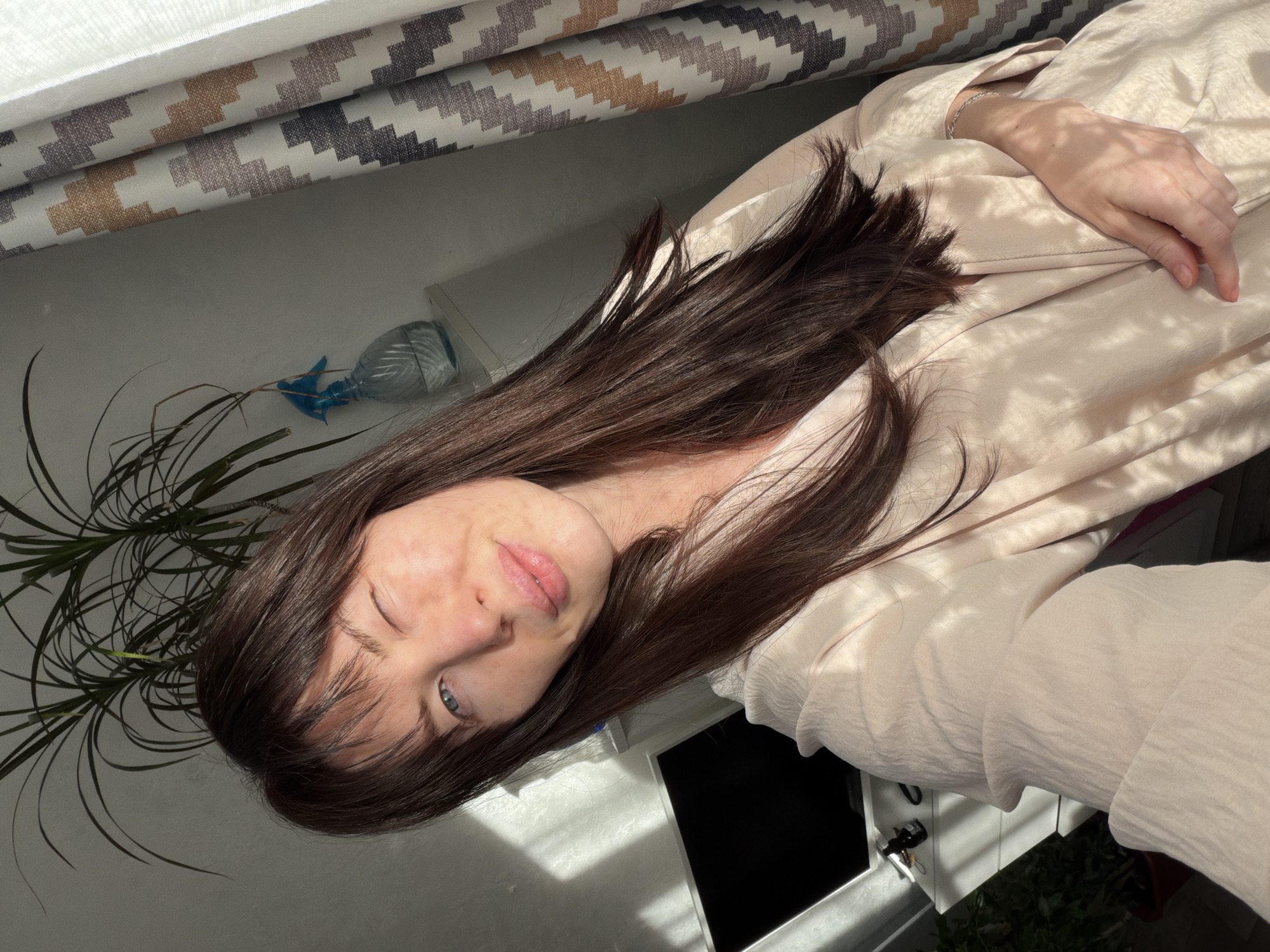 The image shows a woman indoors, possibly in a residence. The woman has dark brown hair and blue eyes. She is wearing a light pink top. Her hand is visible and she wears a bracelet. A plant is visible in the lower left corner, and a patterned curtain is in the background. There is a spray bottle on the windowsill. The location is inferred to be Perm, Russia, though no specific landmarks are visible.